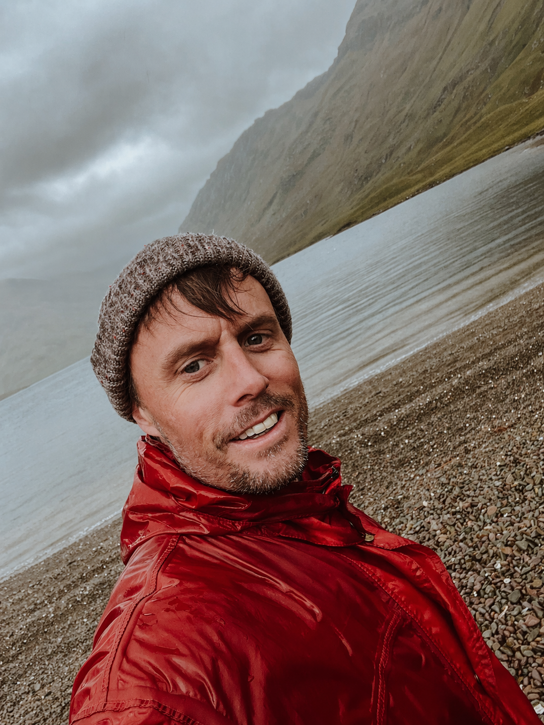A man taking a selfie on a pebble beach with a mountain and cloudy sky in the background, wearing a red jacket and gray beanie.