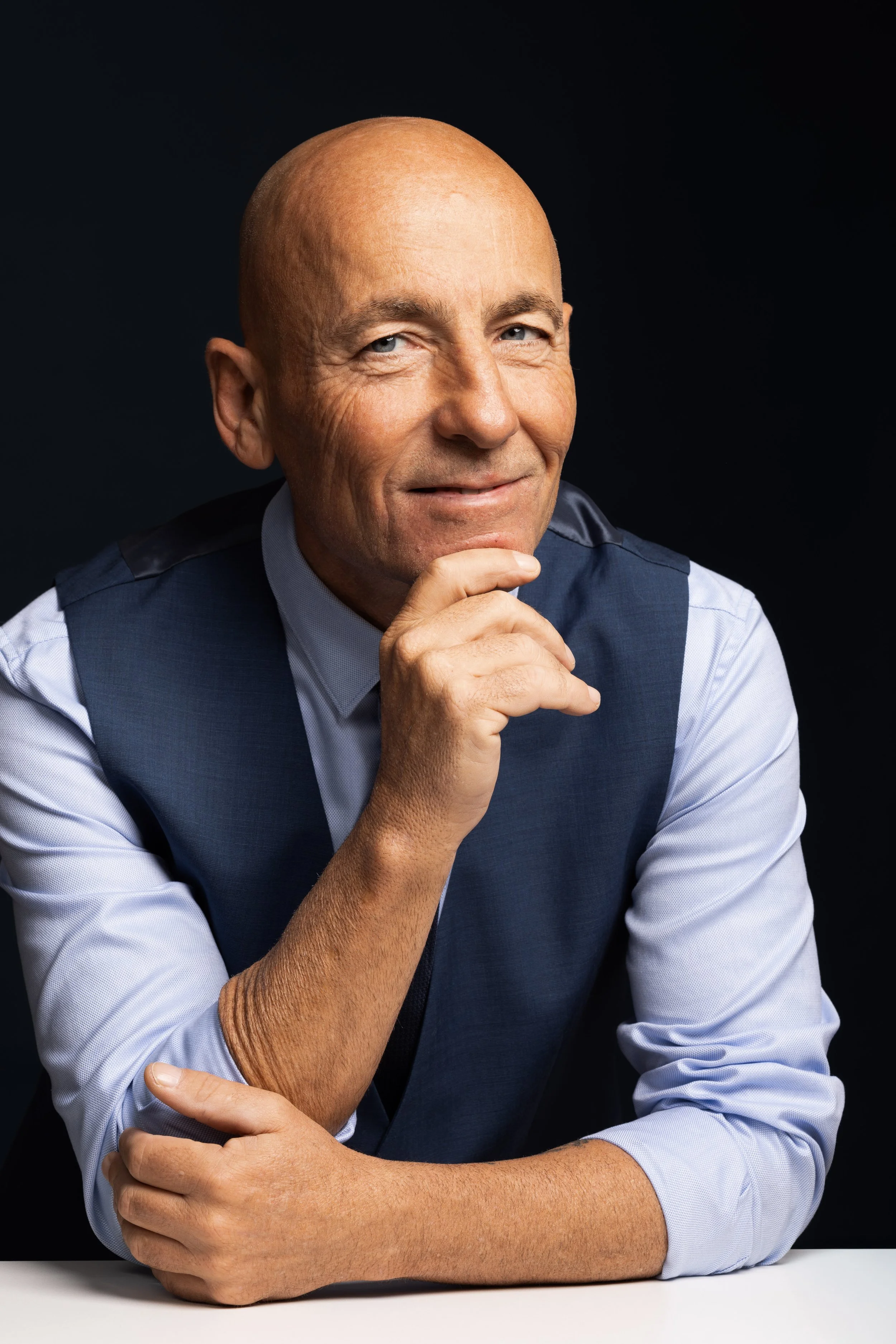 A bald man in a blue vest over a light blue dress shirt, resting his chin on his hand and smiling softly, seated against a dark background.