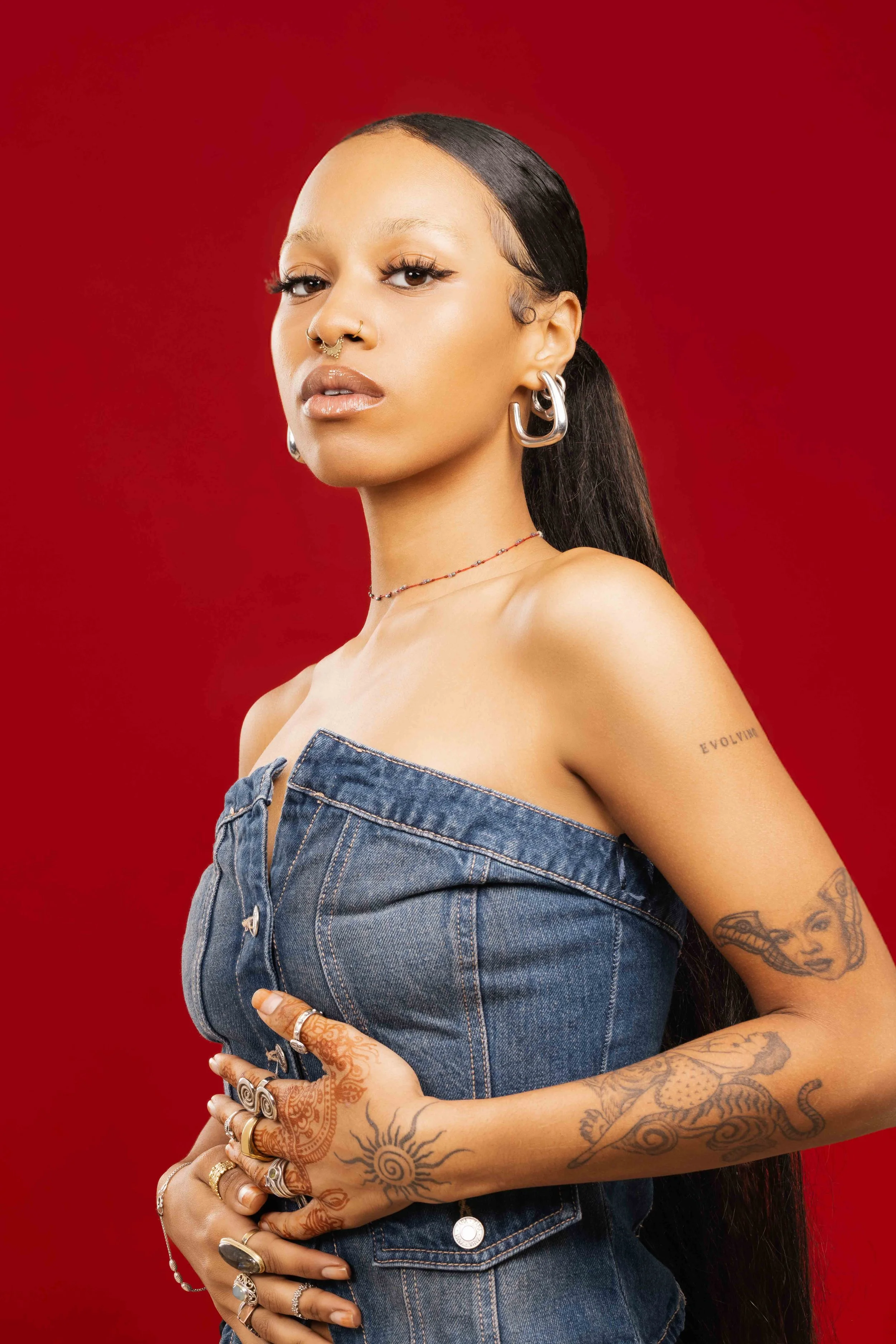 A young woman with long black hair, multiple tattoos on her arms, wearing large hoop earrings, a nose ring, and rings on her fingers, dressed in a denim top, standing against a red background.