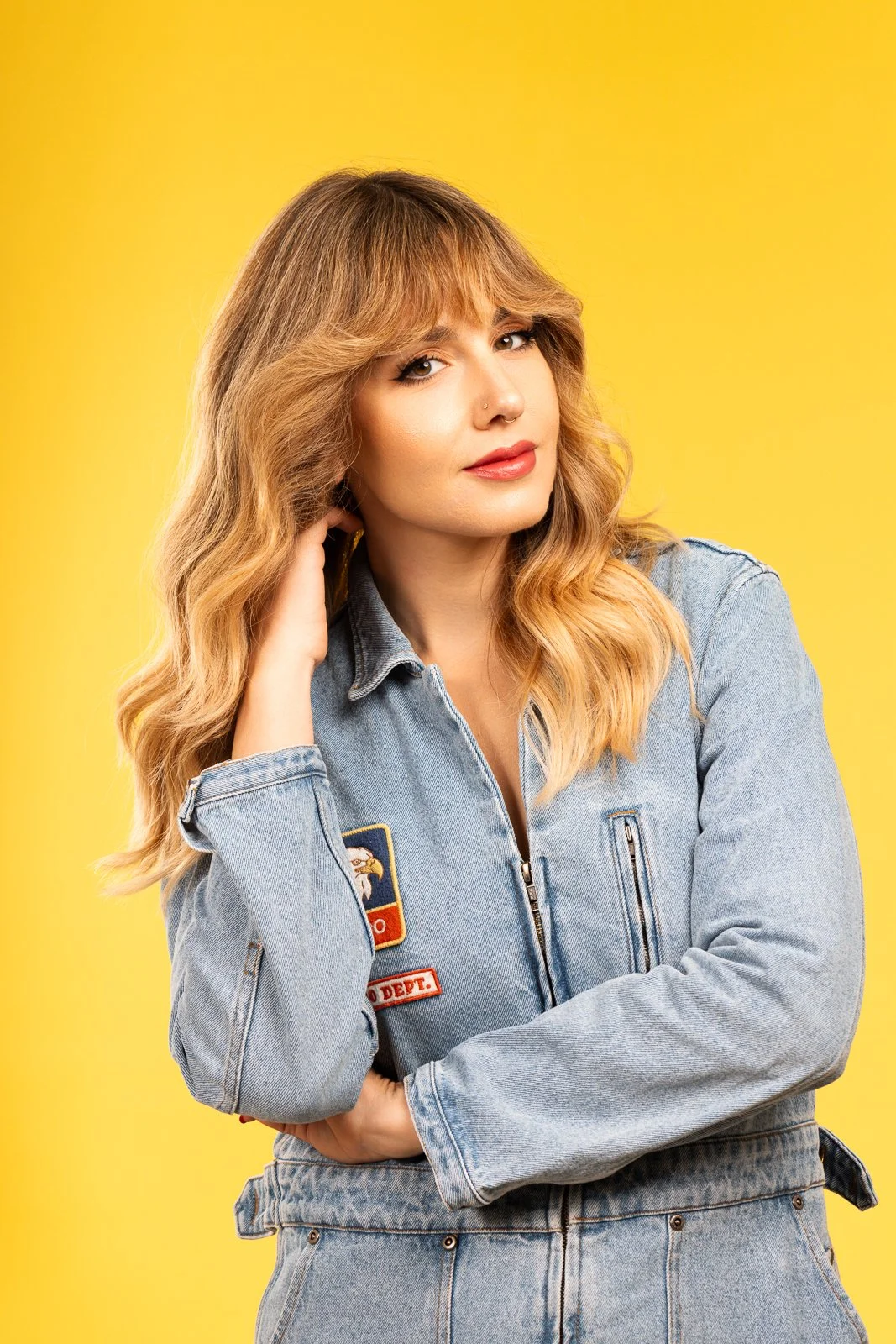 A woman with wavy blonde hair and light makeup, wearing a blue denim jacket with patches, standing against a yellow background.