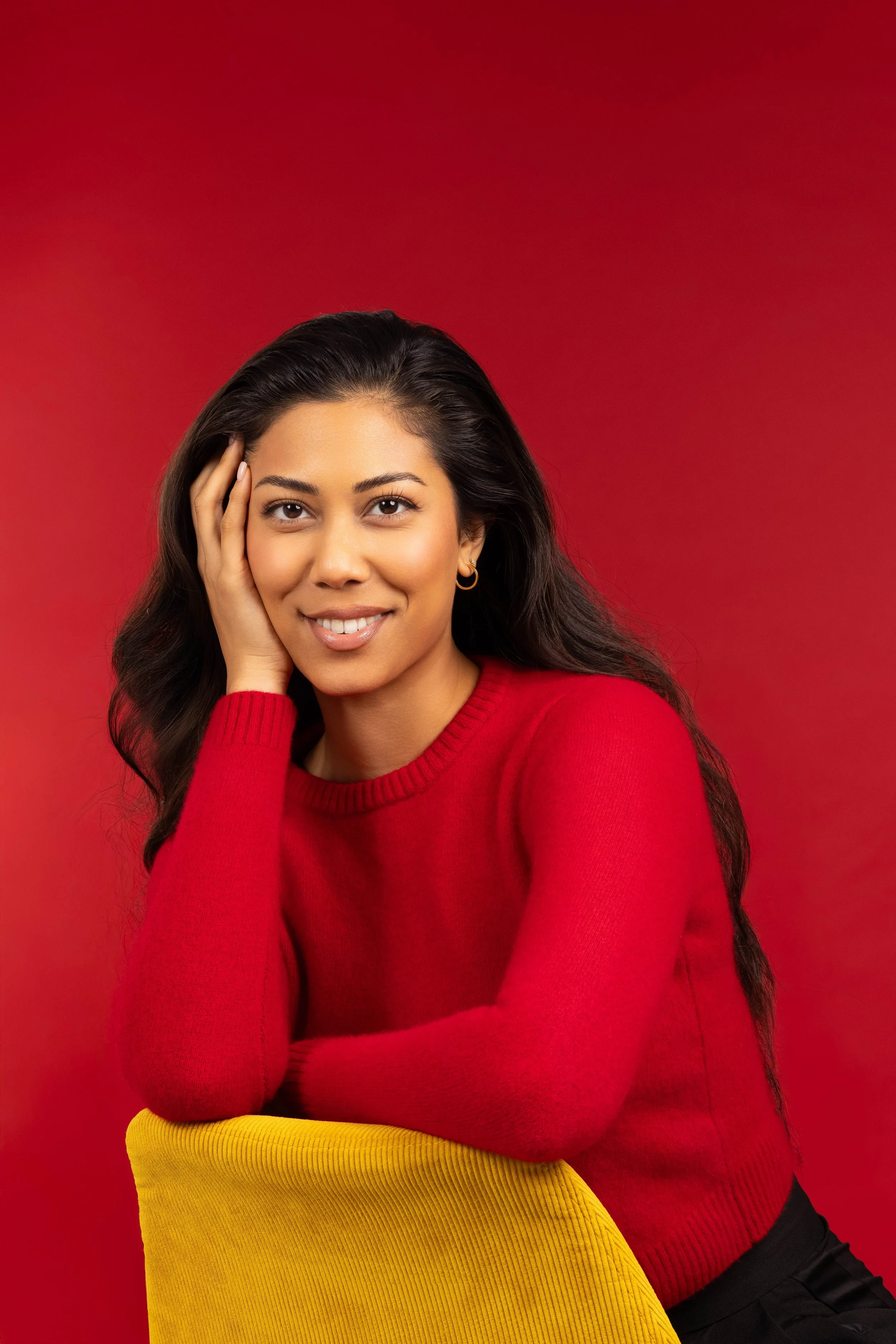 A young woman with long dark hair smiling, wearing a red sweater and gold hoop earrings, sitting against a red background, with a yellow cushion in front of her.