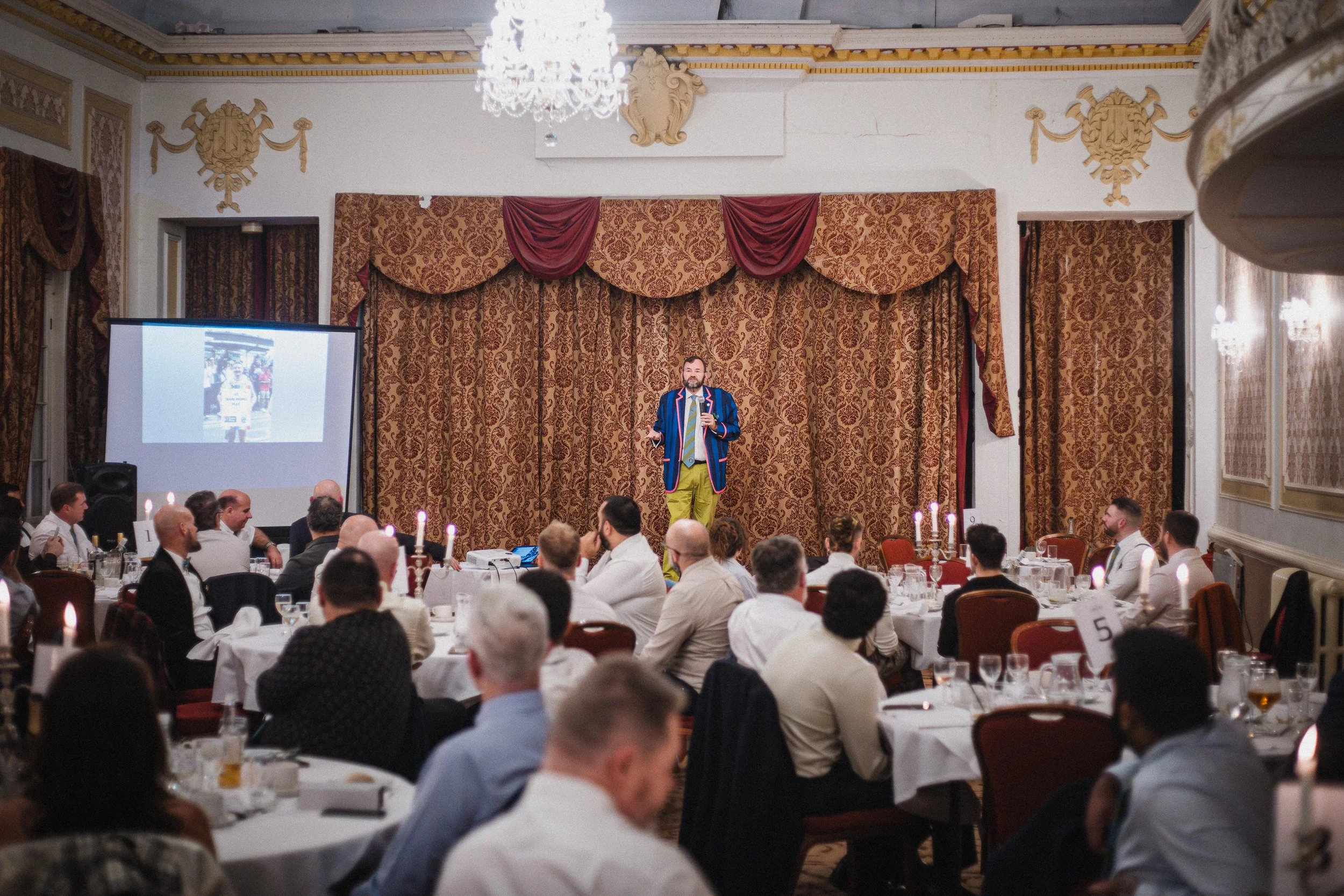 Man dressed in a colorful jacket and yellow pants giving a presentation on stage at a formal gathering or event, with seated audience members at decorated tables.