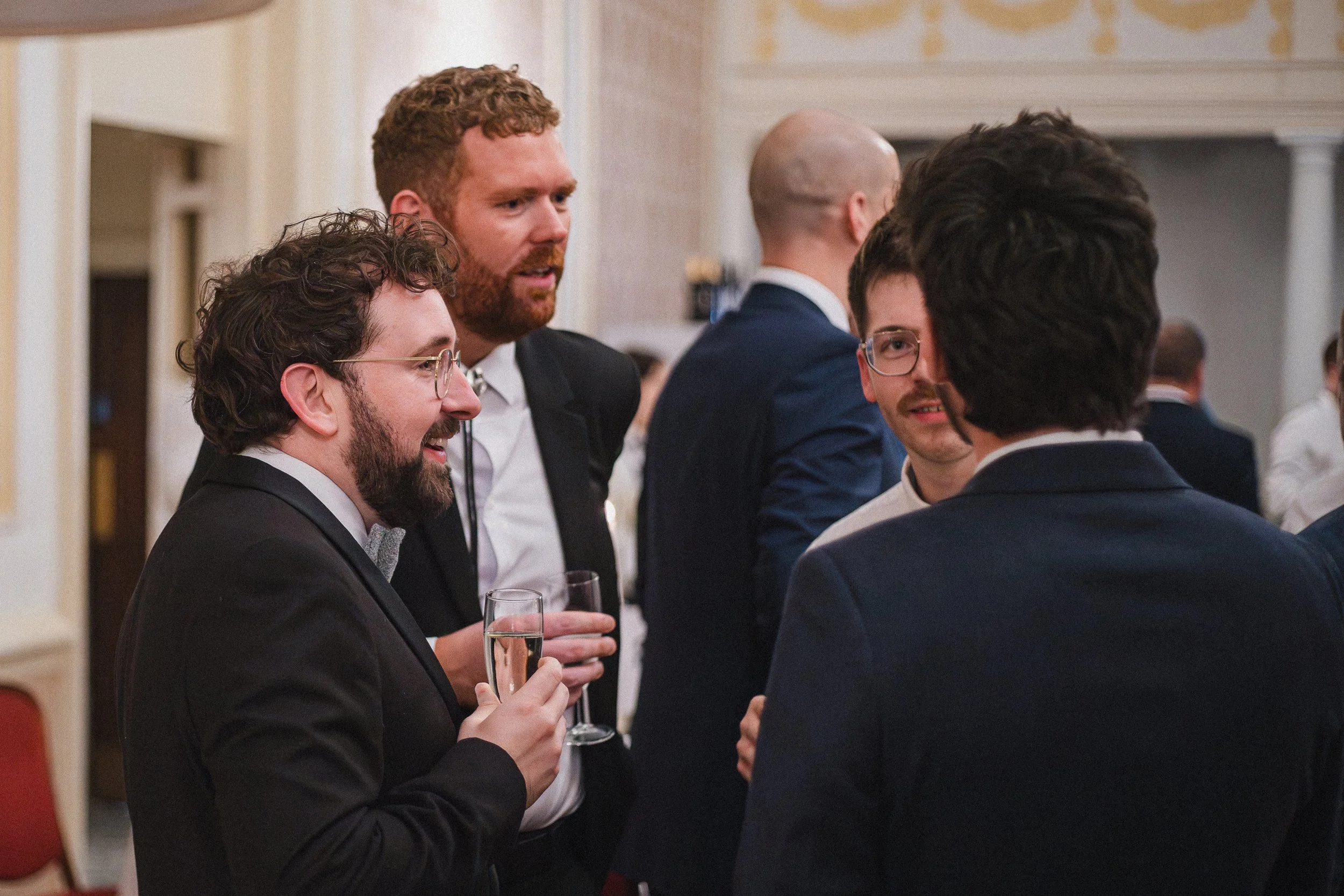 A group of men in suits and tuxedos chatting at a formal event, some holding champagne glasses in a decorated room.