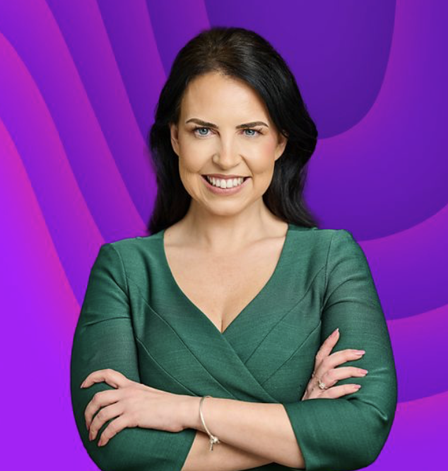 A woman with dark hair, wearing a green dress, stands against a colorful purple and pink abstract background, smiling with crossed arms.