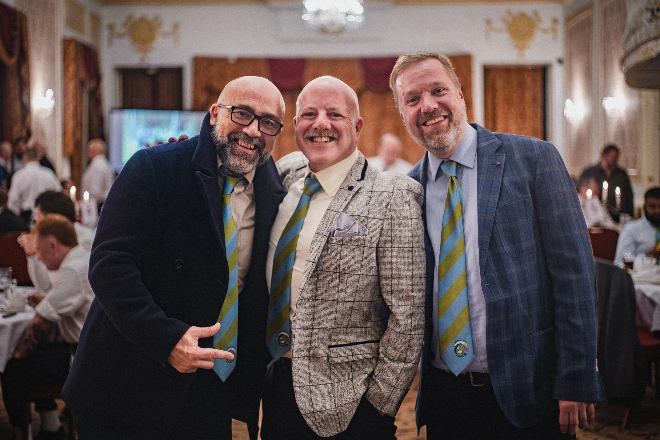 Three smiling men in suits and ties posing together in a formal event hall with other guests in the background.