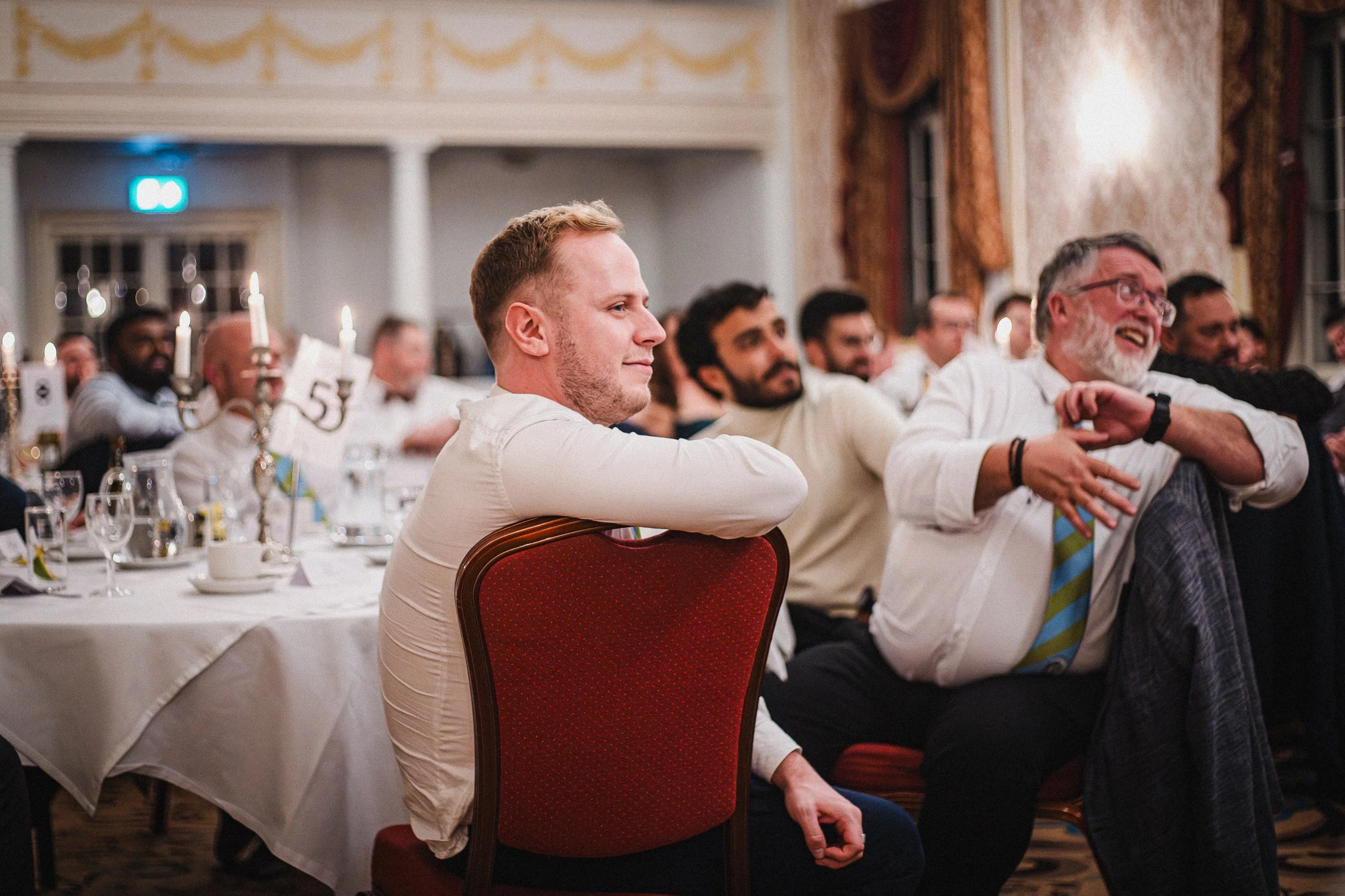 Guests sitting at wedding reception, some smiling and others laughing, in an ornate room with curtains and decorated walls.