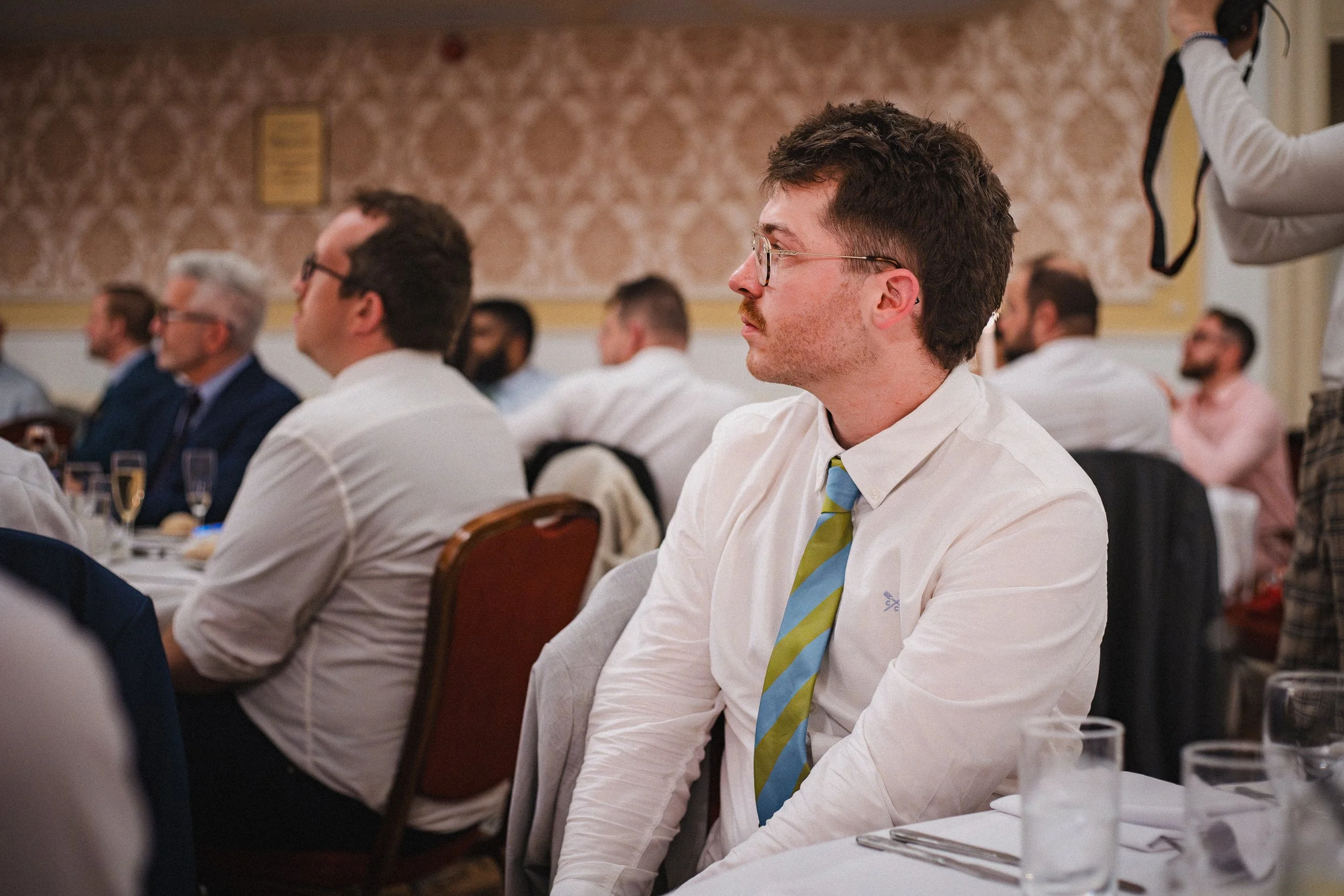 Man with glasses wearing a white dress shirt and a blue and green striped tie sitting at a formal event or conference, with other attendees visible in the background.