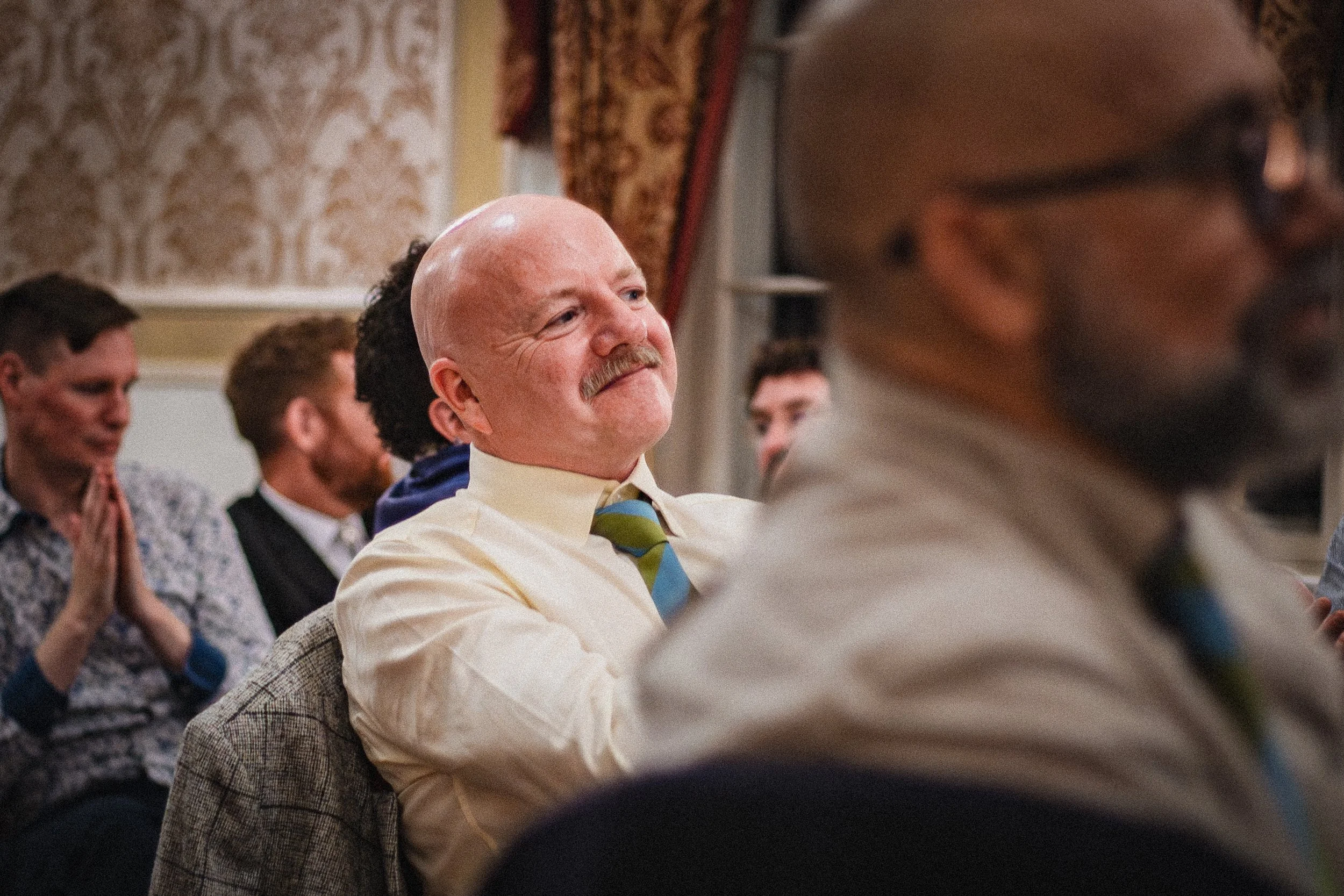 A group of people sitting in an ornate room, with one man in a yellow shirt and striped tie smiling and looking attentive. Other people are visible in the background.