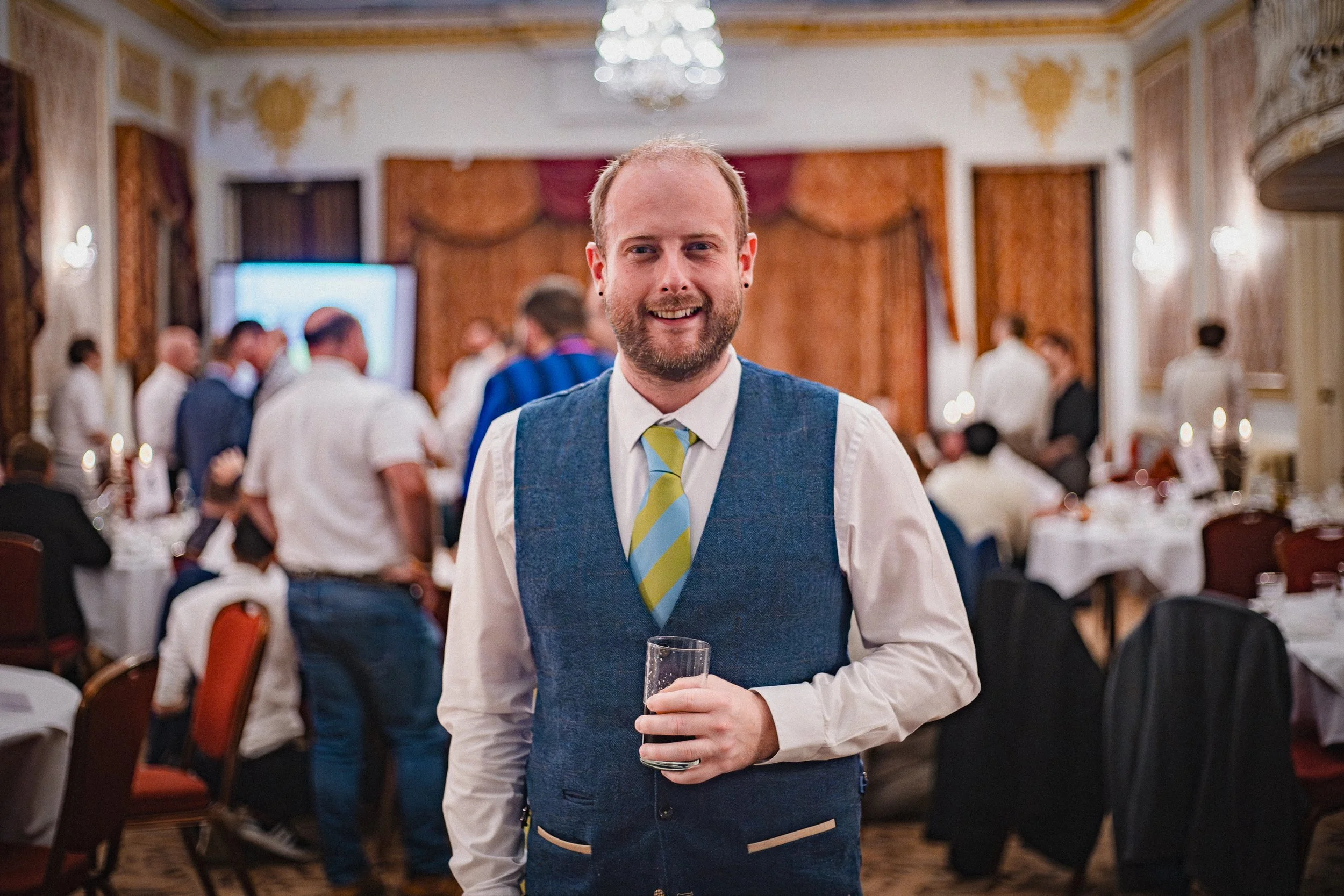 A smiling man in a blue vest, white shirt, and striped green and blue tie holding a glass, at a formal gathering in an ornate room with gold accents and tables with white tablecloths.