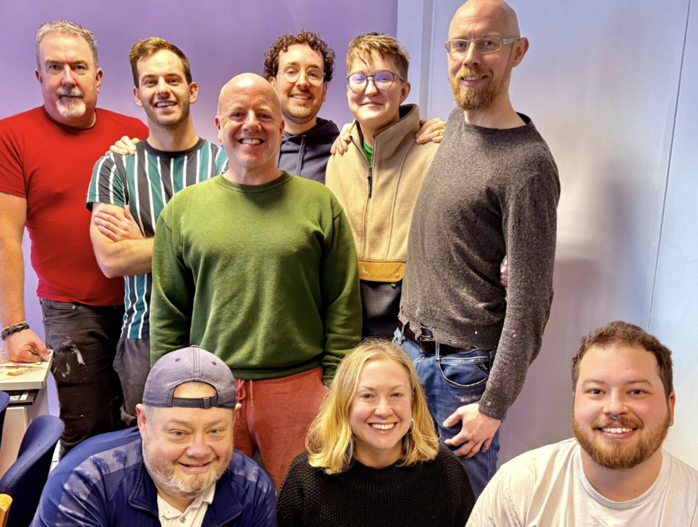Group of nine smiling people posing together in an indoor setting.