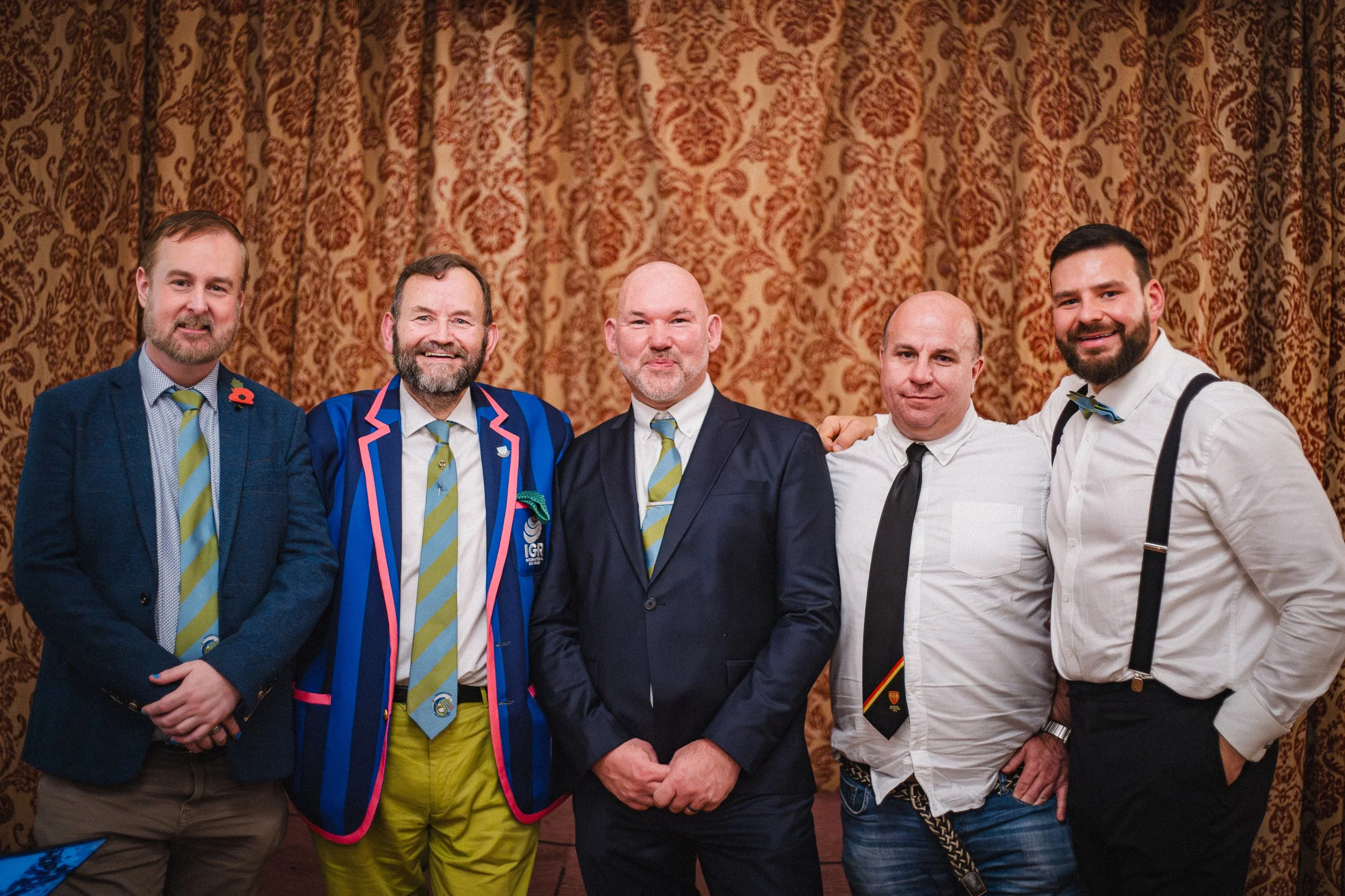 Five men dressed in suits and shirts, standing in front of a patterned curtain, smiling at the camera.