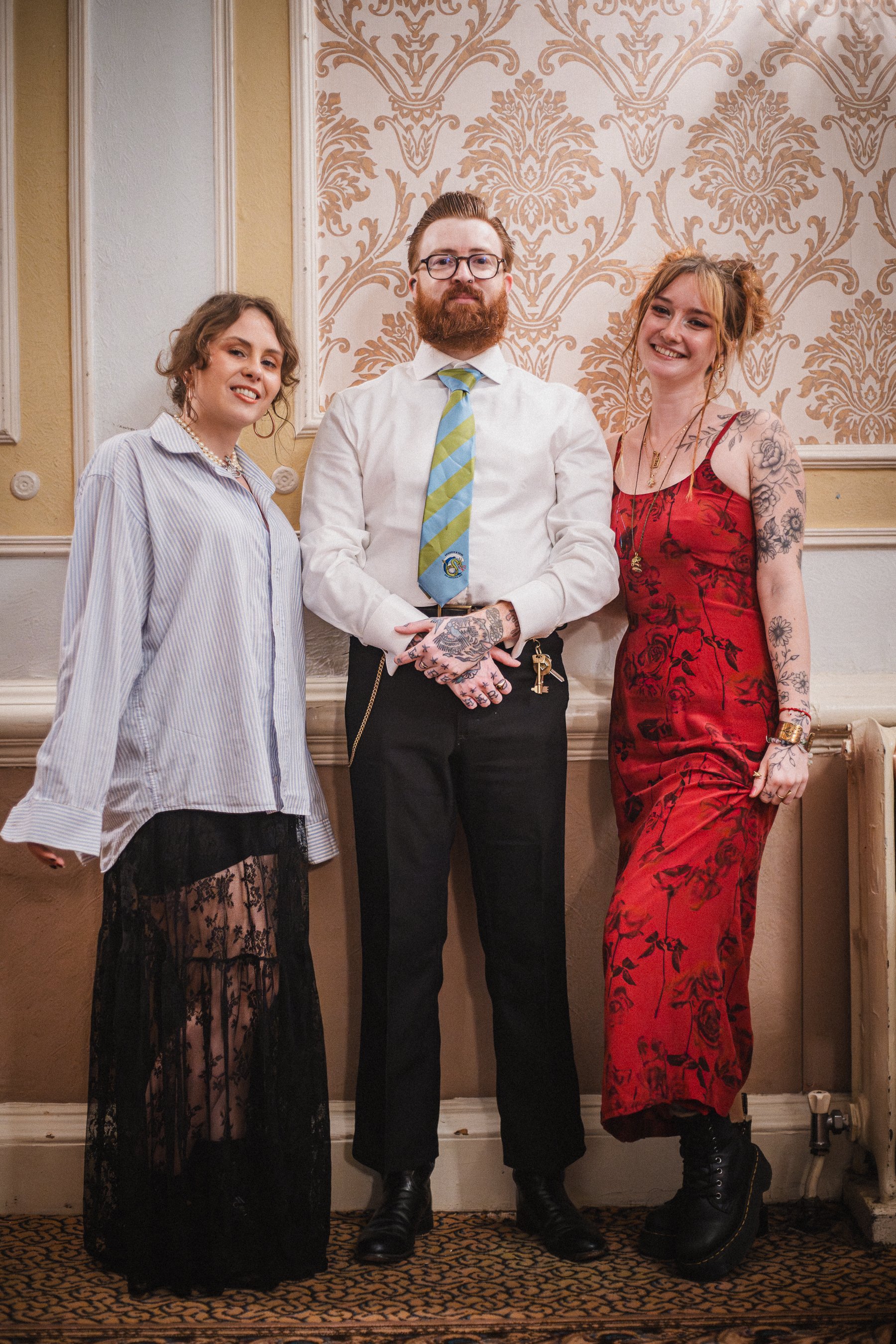 Three people posing together indoors, with ornate wallpaper background. The person on the left is wearing a striped shirt and a long black lace skirt. The person in the middle, a man with glasses and a beard, is dressed in a white shirt, striped tie,