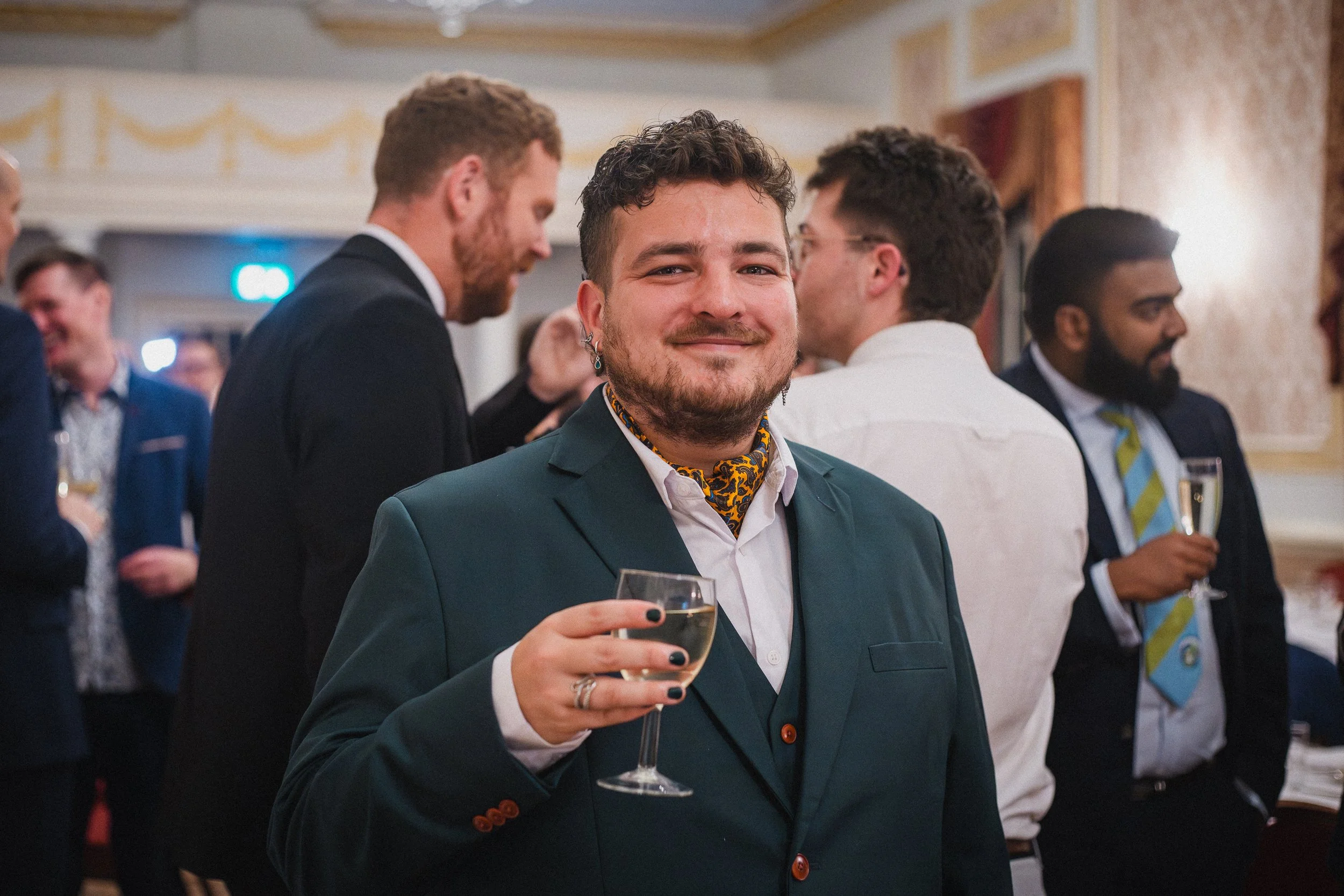 A smiling man dressed in a dark green suit with a patterned scarf around his neck, holding a glass of champagne at a social gathering with other well-dressed people in the background.