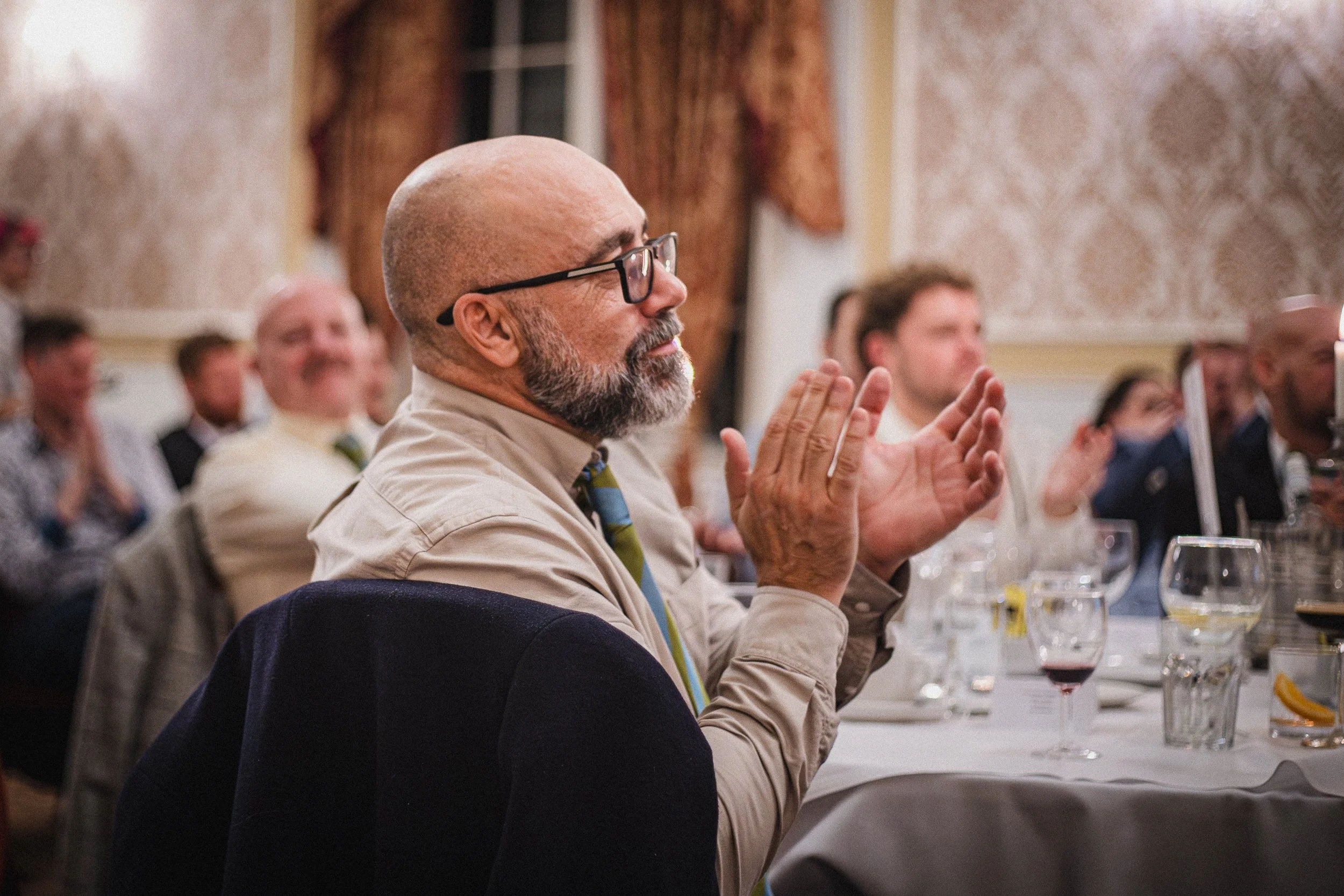 A man with glasses, a bald head, and a beard claps during an event, sitting at a table with wine glasses and other people in the background.