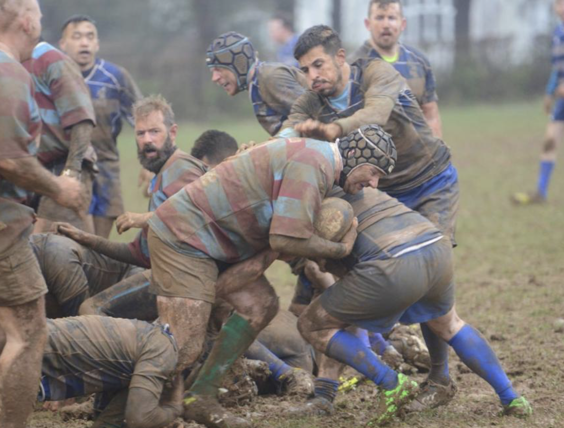 A muddy rugby match with players engaged in a scrum on a grassy field, some wearing helmets and muddy jerseys.