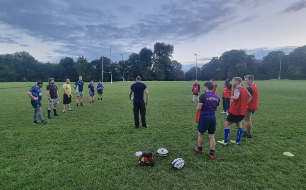 A group of adults and children on a rugby or football field listening to a coach during practice or a training session, with the field and trees in the background at dusk.