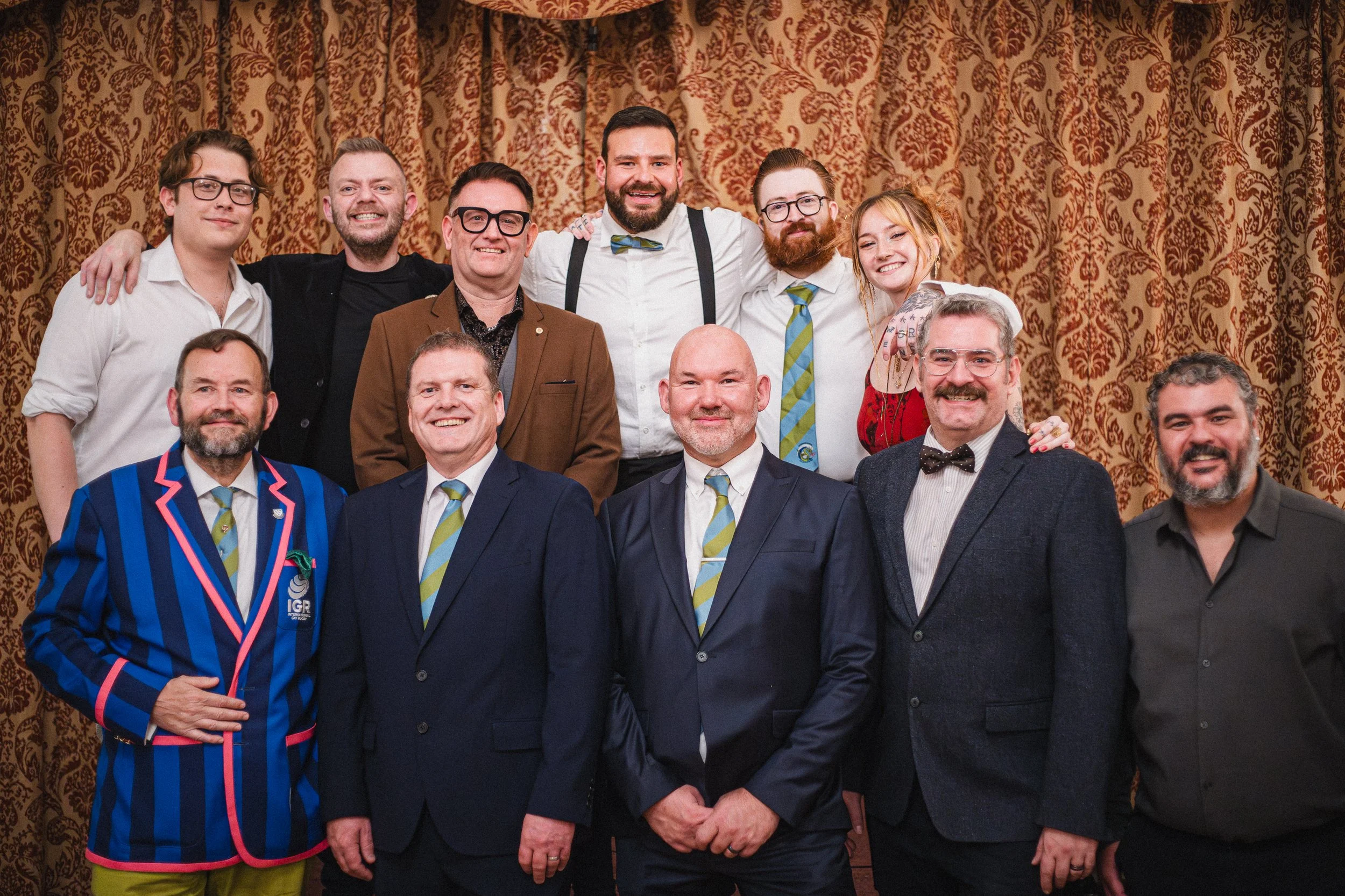 Group of twelve people in formal and semi-formal attire posing in front of a gold-patterned curtain, with some smiling and others with neutral expressions.