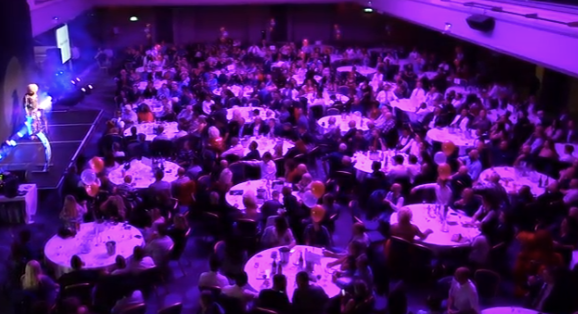 A large banquet hall filled with round tables and many guests, with a stage and performer on the left.