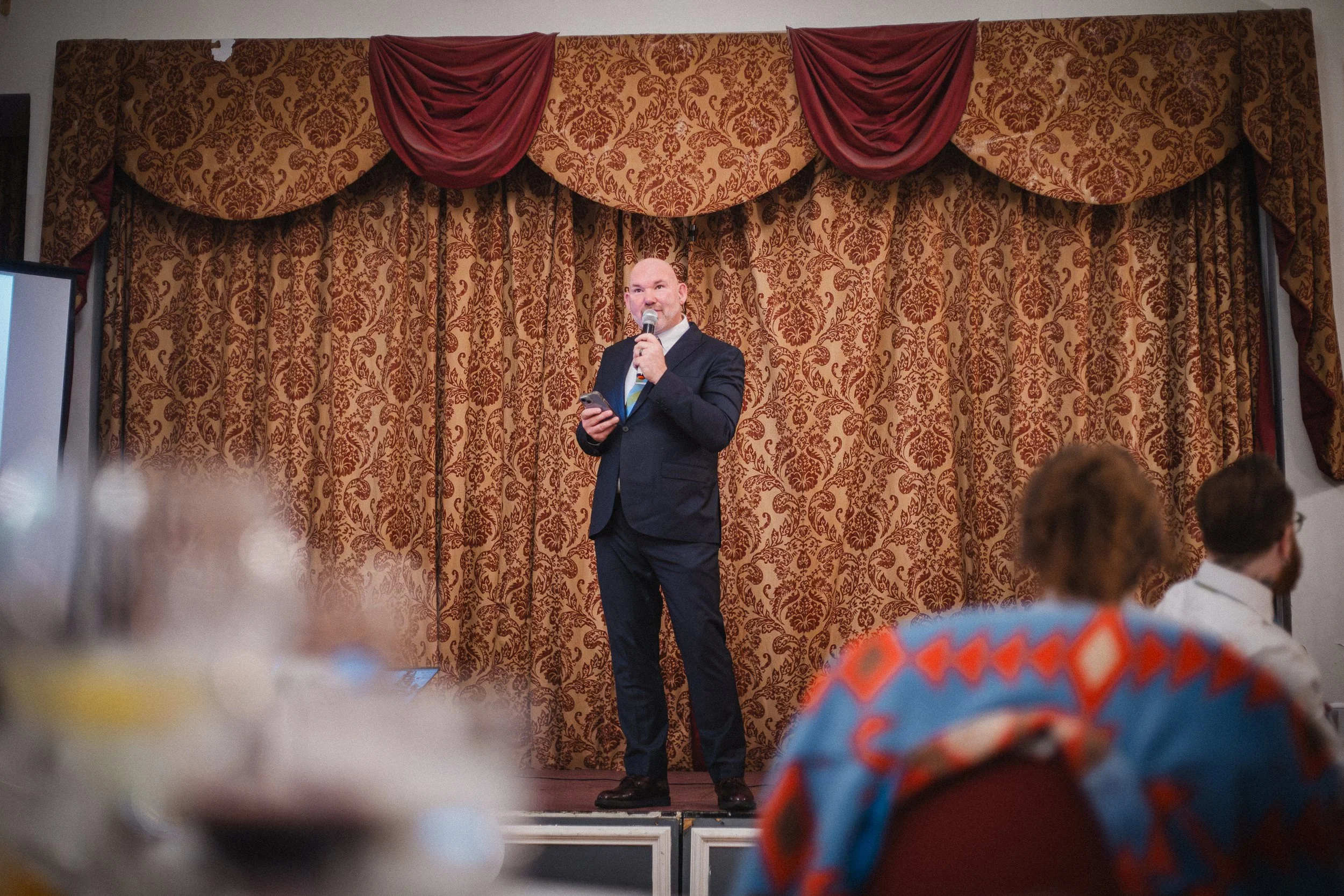A man in a dark suit and tie holding a microphone and reading from a phone on a stage with ornate burgundy and gold curtains.