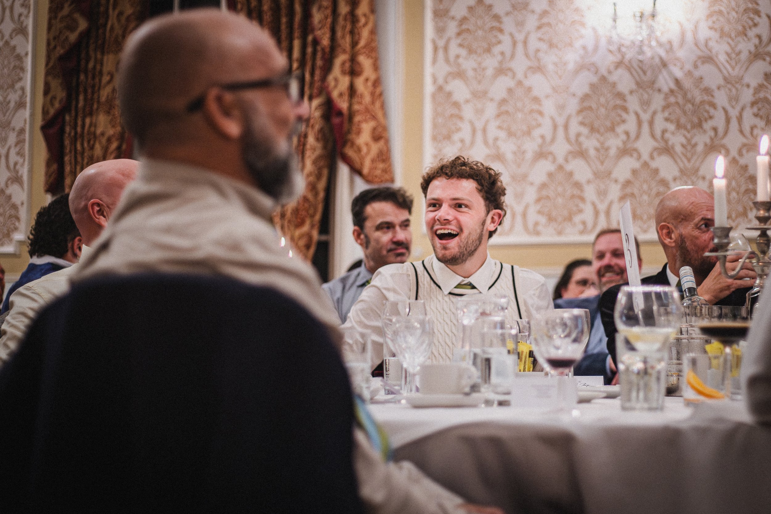 A group of people sitting at a banquet table in a decorated room, with a young man smiling and laughing at the center, surrounded by others engaged in conversation.