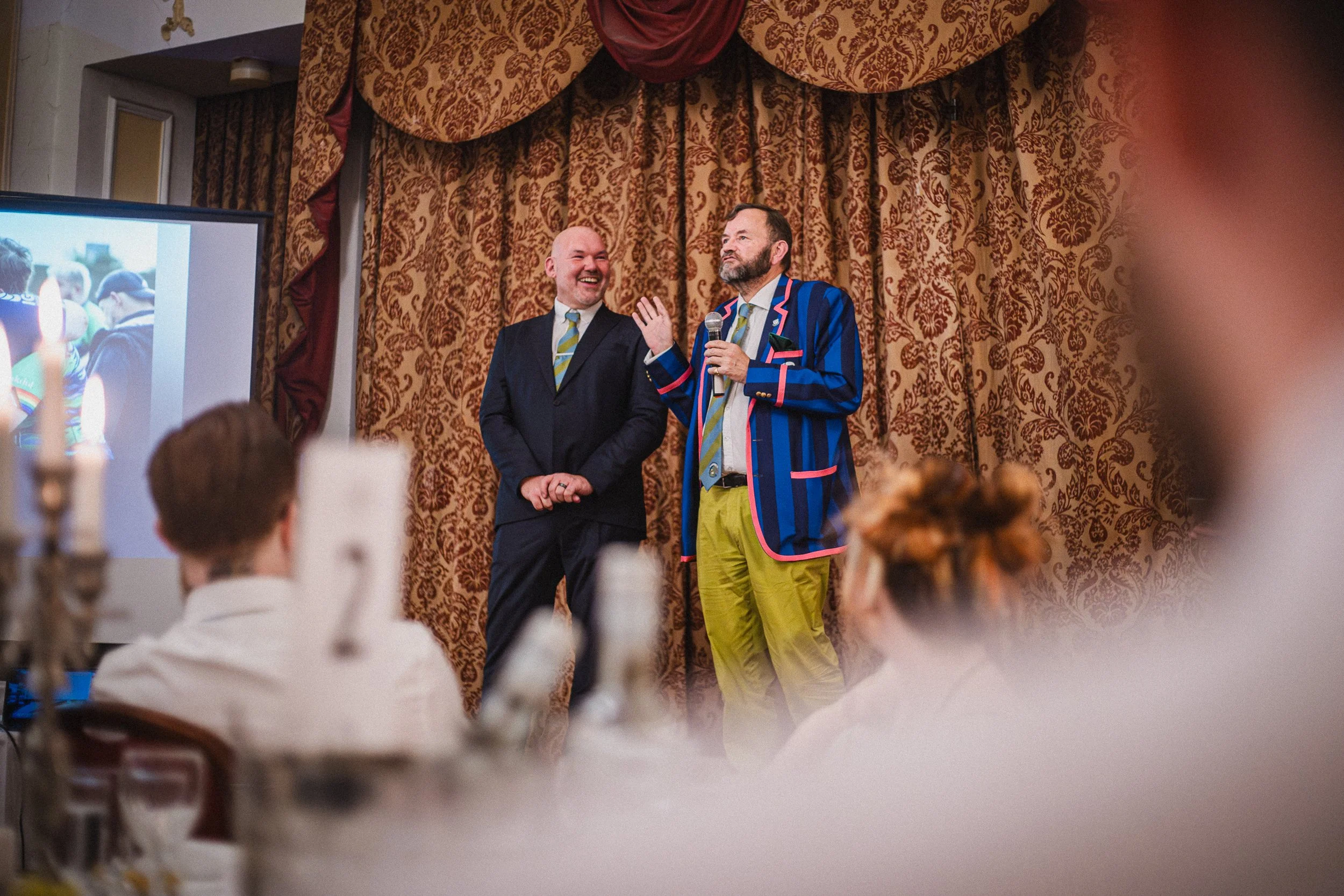 Two men in colorful suits speaking at an event, one holding a microphone, with an audience in the foreground and a decorated curtain behind them.