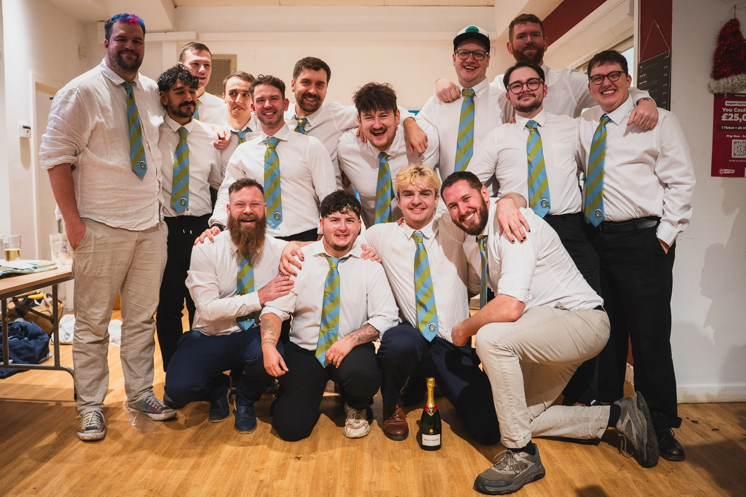 Group of 16 men celebrating together indoors, some wearing white shirts with striped blue and green ties, and others dressed more casually. They are smiling, with some kneeling, sitting, or standing, and one man in front holding a bottle of champagne.