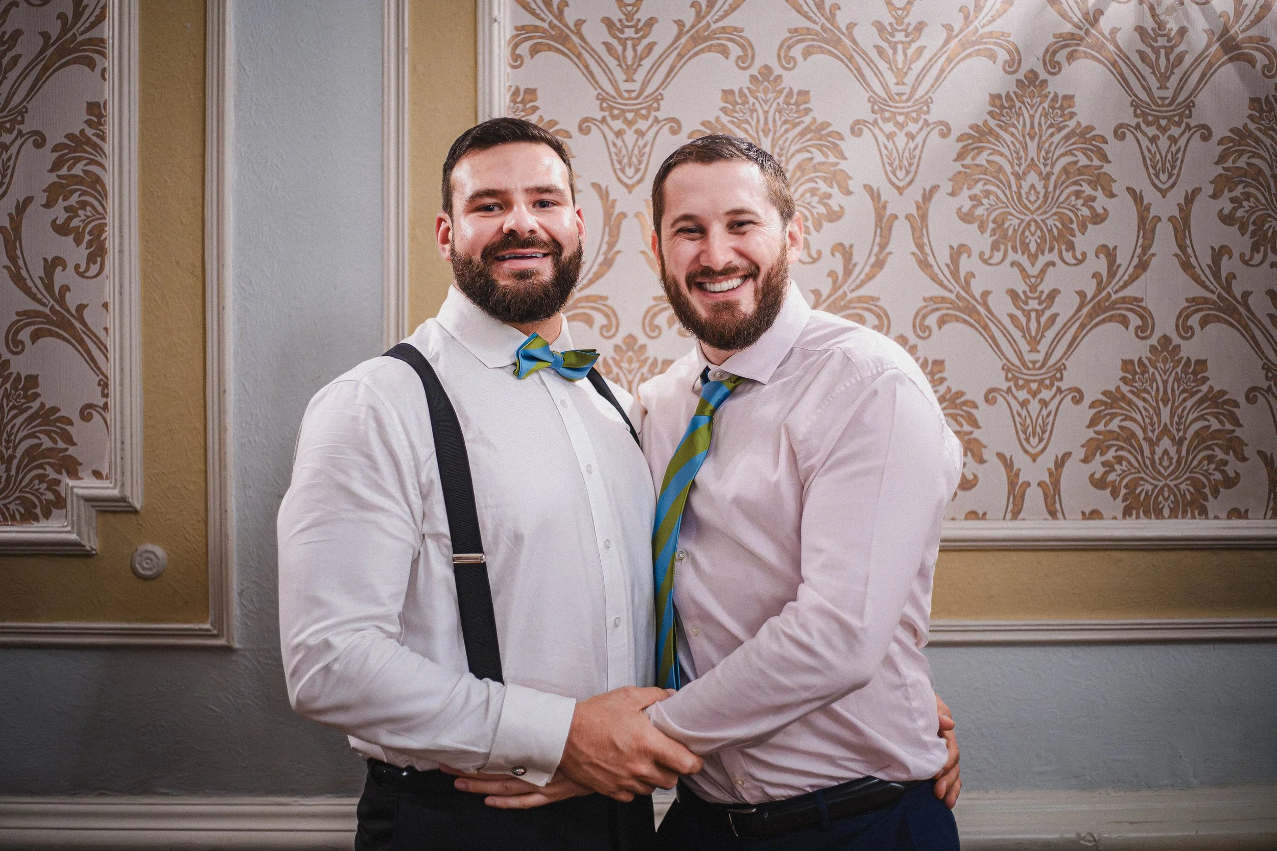 Two men with beards smiling and holding hands at a formal event, standing in front of an ornate patterned wall.