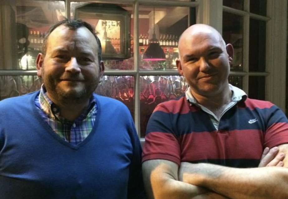 Two men standing side by side in a restaurant, smiling. One is wearing a blue sweater over a plaid shirt, and the other is wearing a red and navy striped polo shirt with crossed arms.