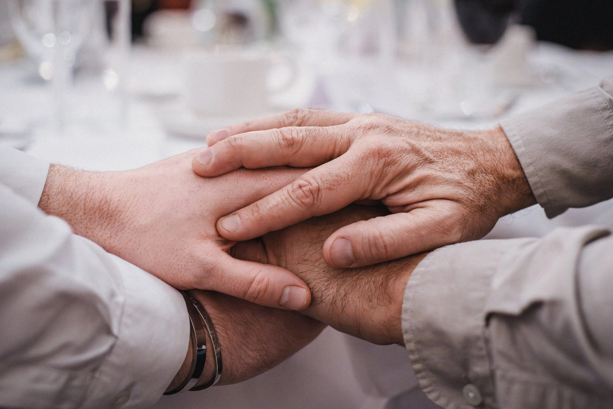 Two pairs of hands holding each other, one pair appears younger and the other older, on a table set for dining.