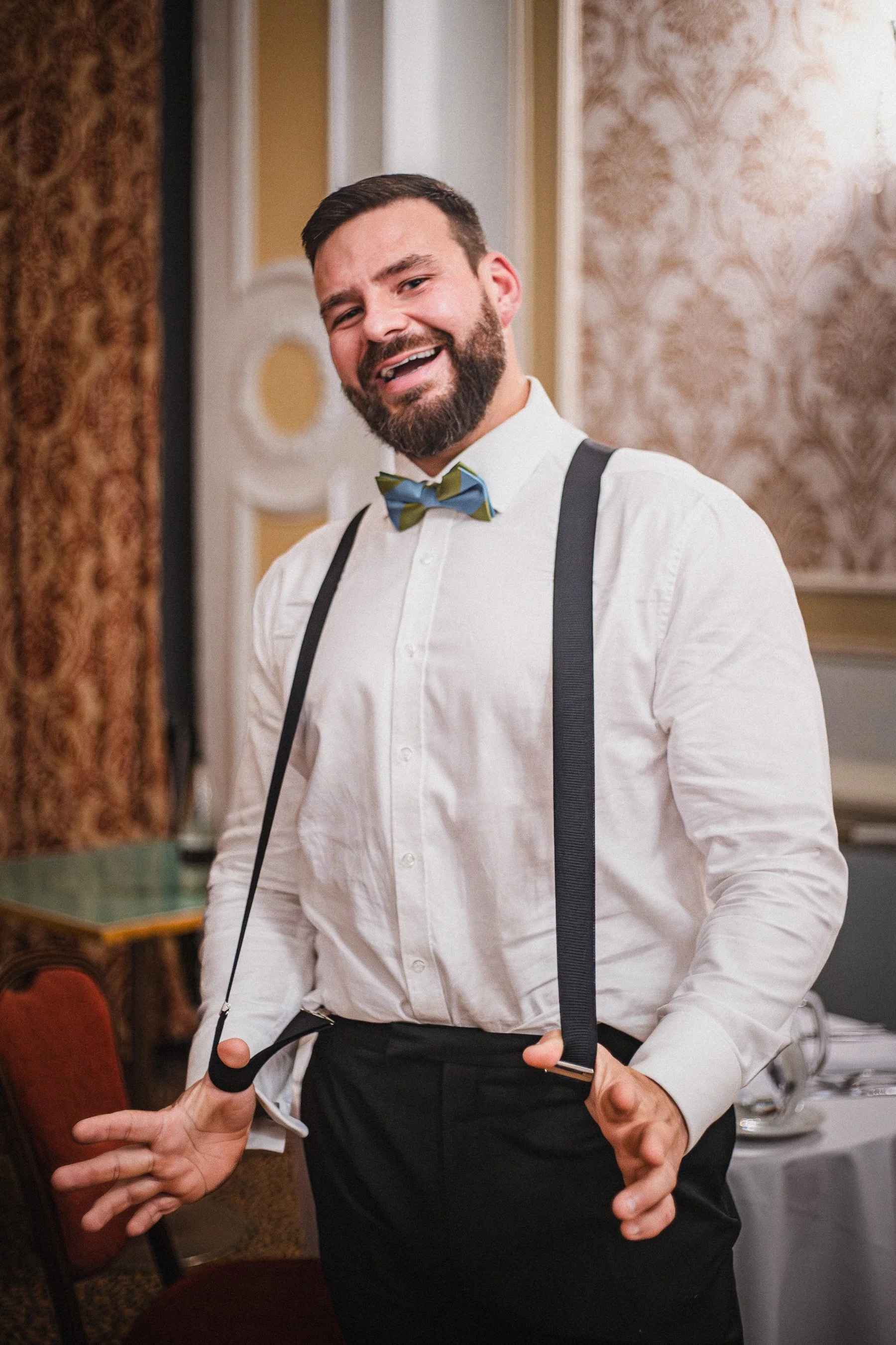 A man with a beard wearing a white shirt, bow tie, and suspenders smiling and posing indoors