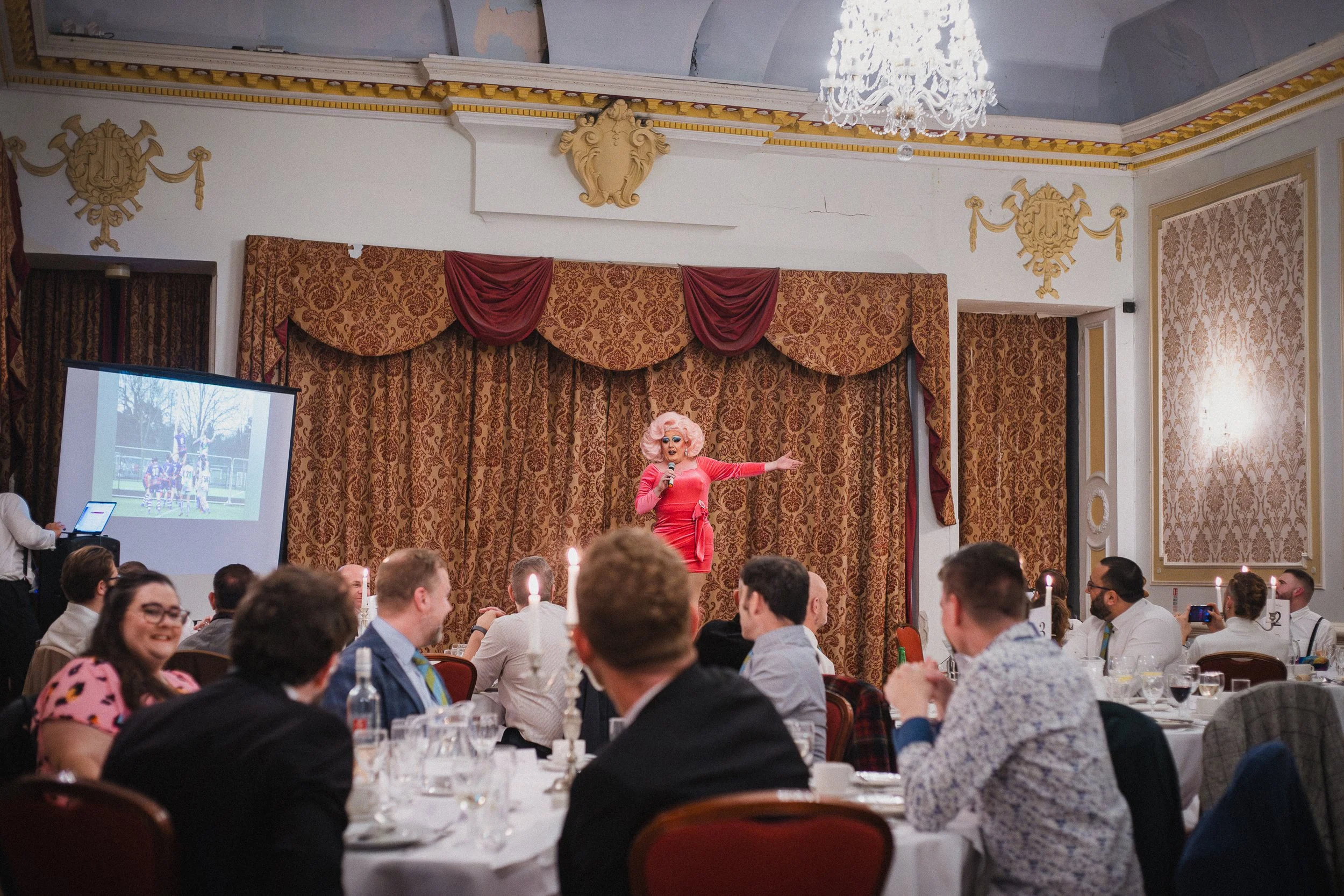A woman dressed as a performer or drag queen, in a bright pink dress and large pink wig, is on stage holding a microphone, in a banquet hall with an audience seated at tables. The hall has ornate decor, chandeliers, and a large curtain backdrop.