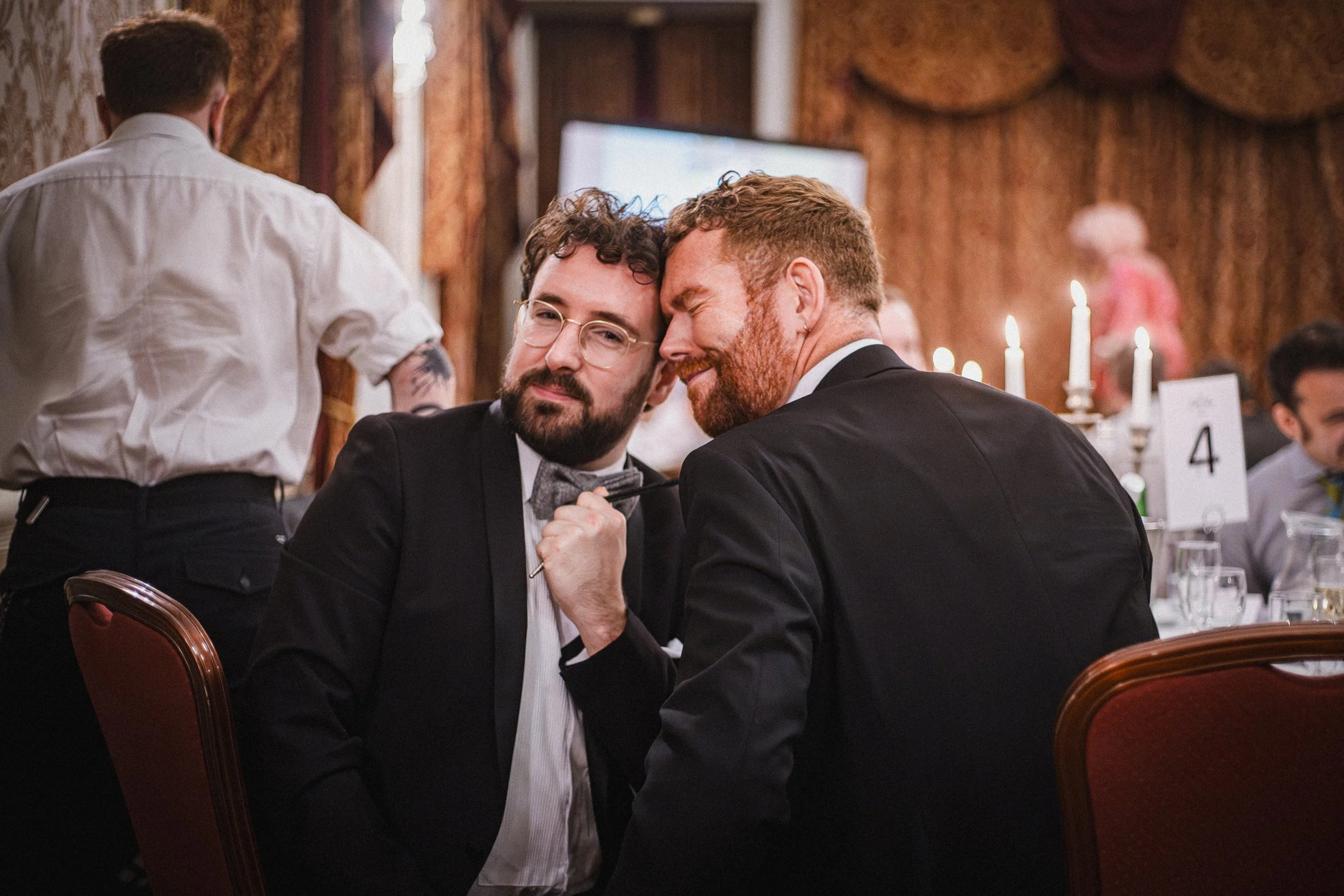 Two men in suits, one with glasses and a gray bow tie, the other with a red beard, sitting close together at a formal event in a decorated room with lit candles.