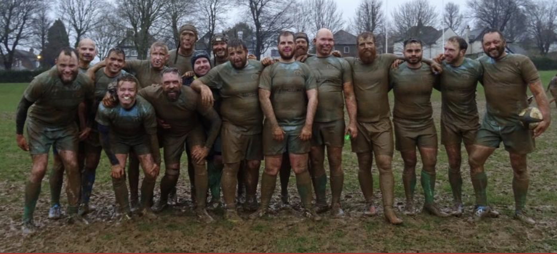 A group of men after a muddy rugby game, smiling and posing outdoors.