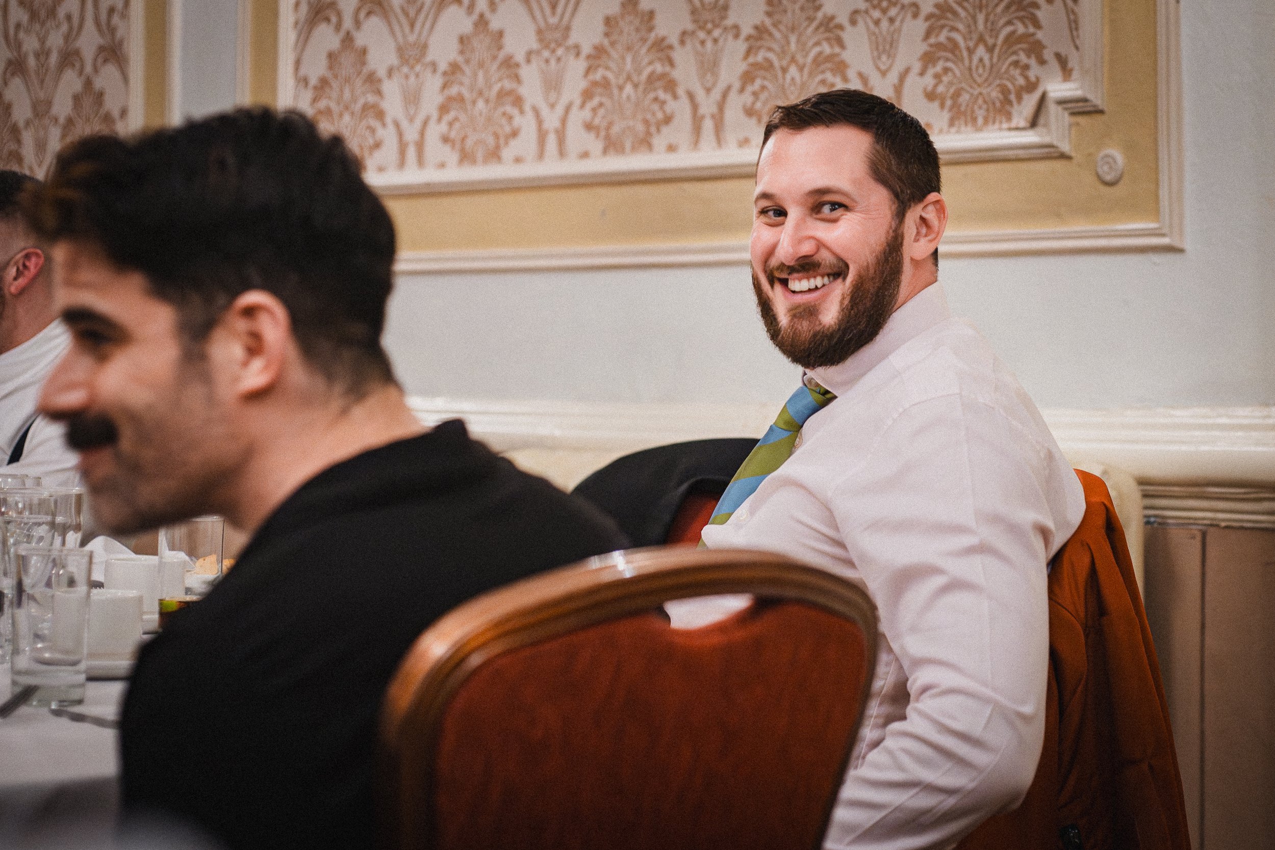 A man with a beard and short hair smiling while sitting at a table during a formal event or dinner. He is wearing a white shirt and a colorful striped tie, with a brown jacket draped over his chair. The background shows ornate wallpaper and a decorat