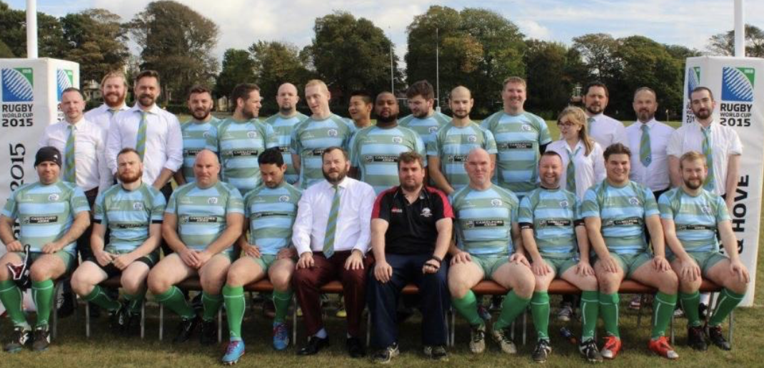 Photograph of rugby players and team staff posing for a team photo outdoors during daytime. The players are dressed in teal and black striped jerseys, shorts, and socks, with some wearing rugby patches and gear. The background features trees and rugby goalposts, with banners indicating a 2015 rugby World Cup event.