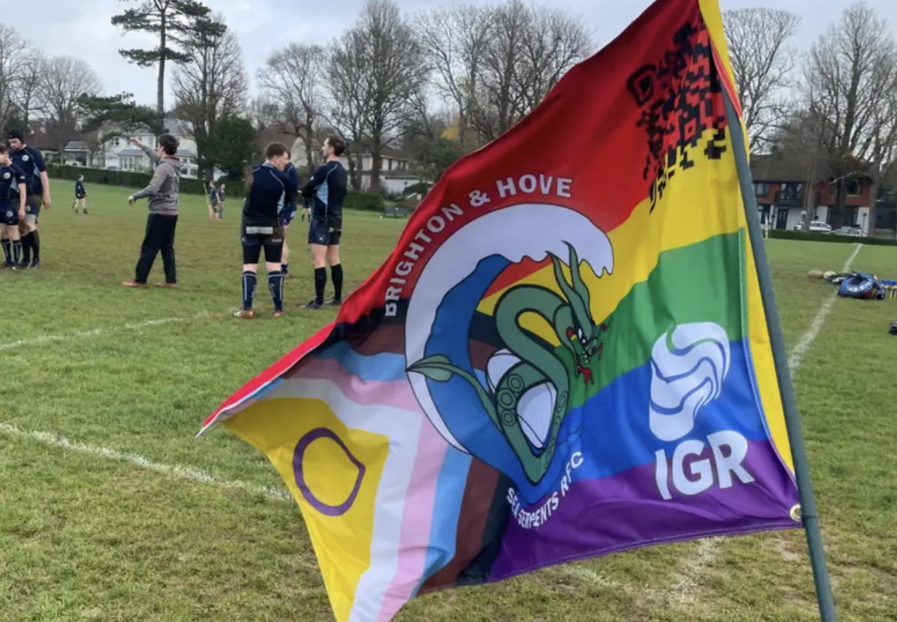 A colorful rainbow-themed flag featuring a green dragon, the text 'Brighton & Hove', and additional letters 'IGR' on a grassy rugby field with players and coaches in the background.