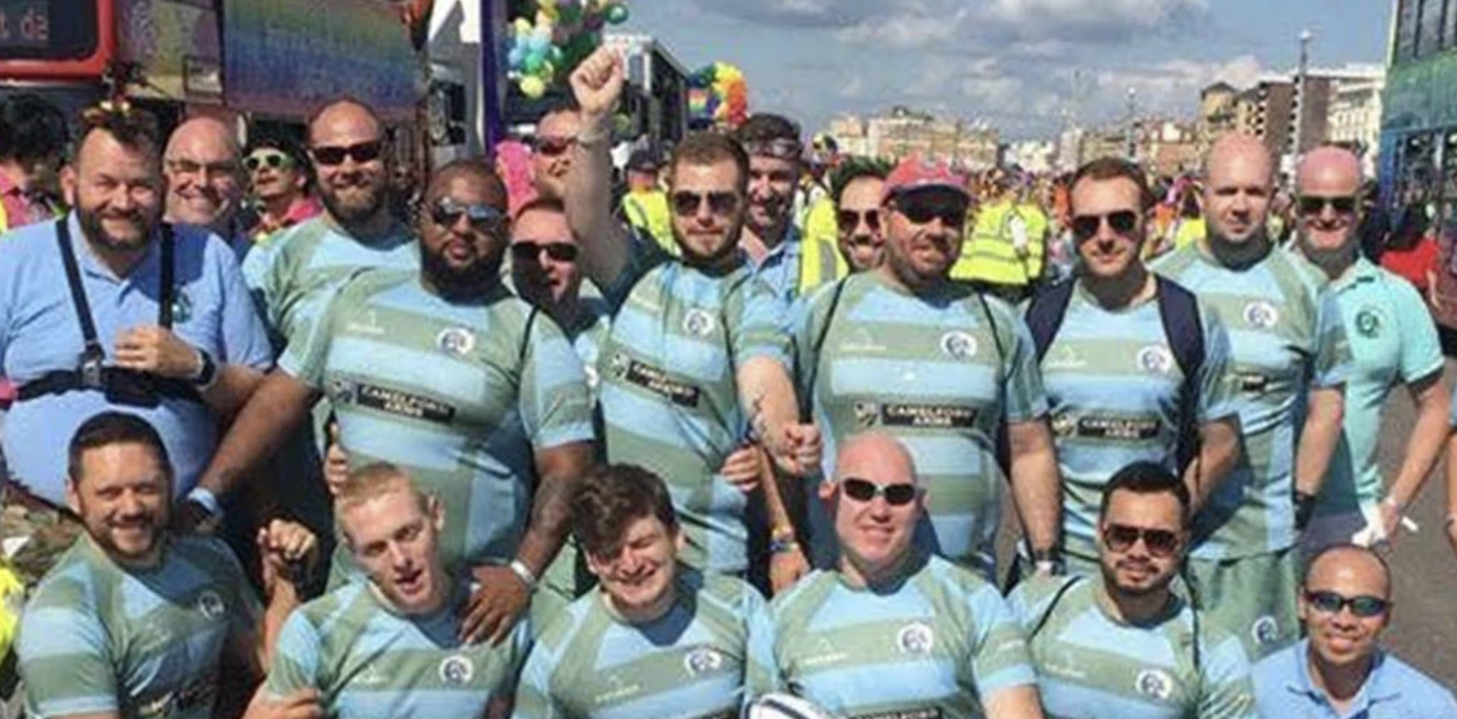 A group of smiling men in matching striped shirts posing for a photo in a crowded outdoor event, with some wearing sunglasses and backpacks, and a cityscape in the background.