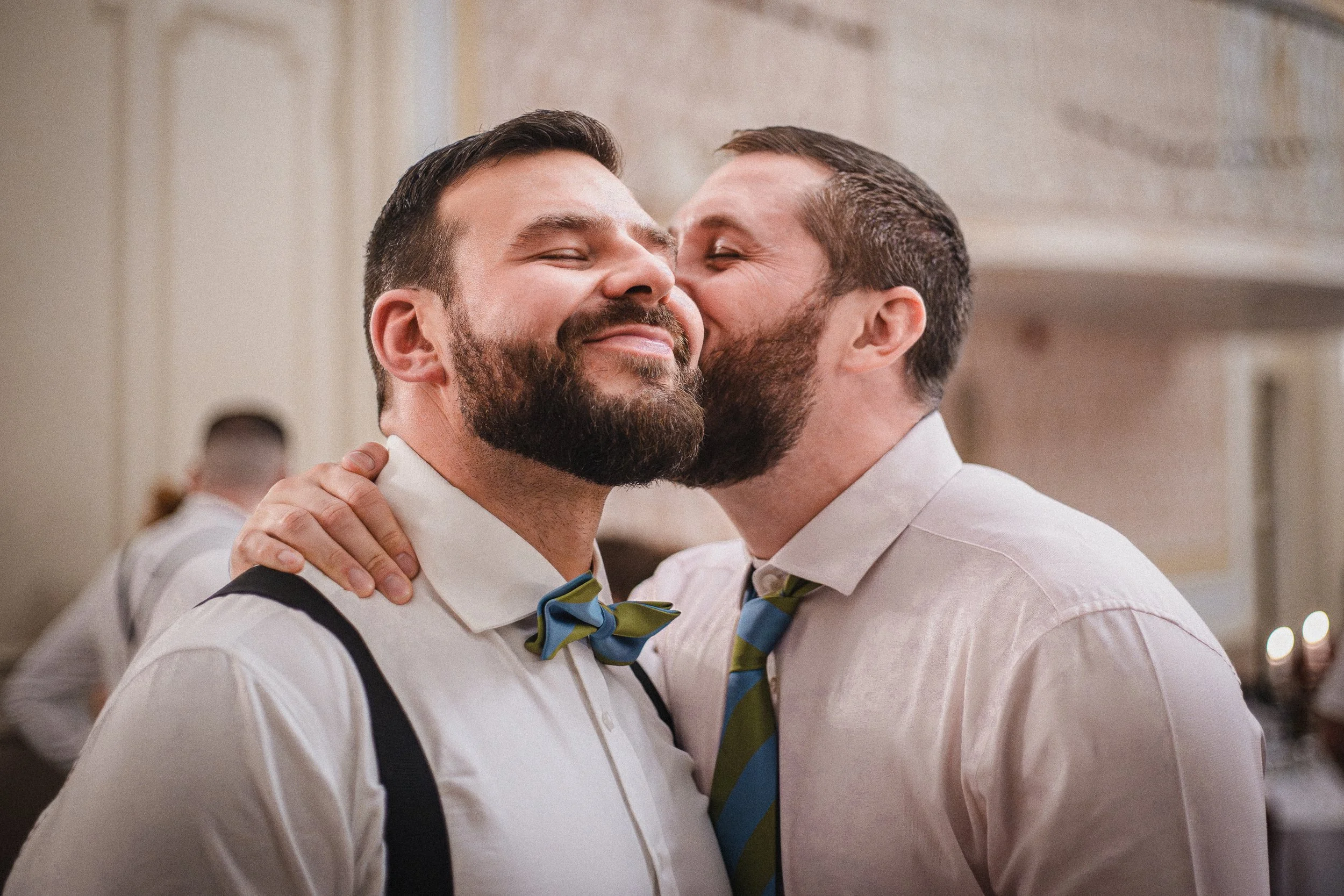 Two men with beards and short hair sharing a kiss on the cheek during a celebration. One man is smiling with eyes closed, while the other is kissing him on the cheek. They are dressed in formal attire, with one wearing suspenders and a bow tie, and t