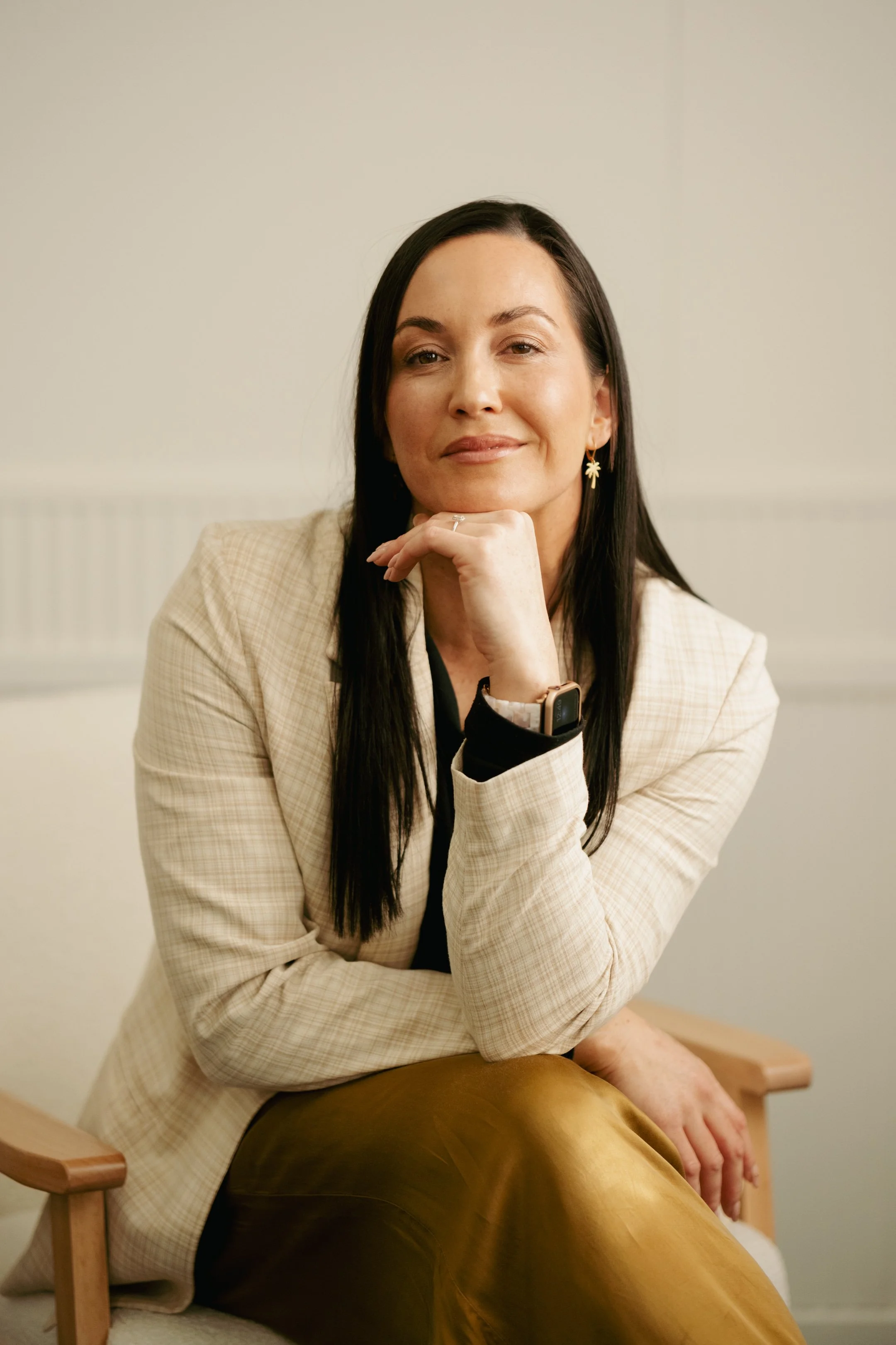 A woman with dark hair, wearing a beige blazer and gold pants, sitting on a wooden chair, resting her chin on her hand and smiling at the camera.