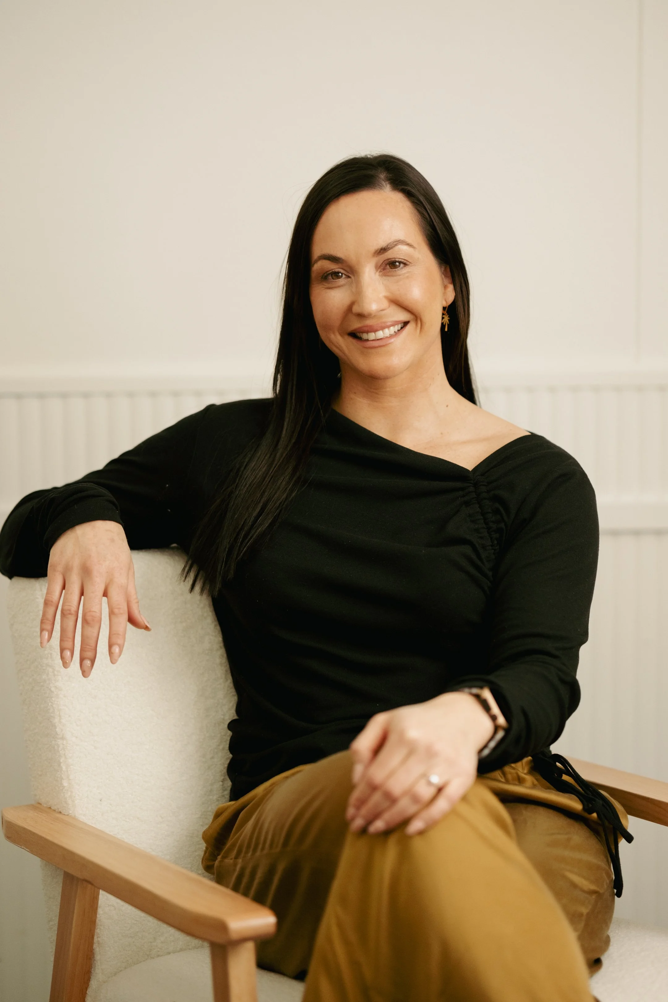 A woman sitting on a wooden chair with a cream-colored upholstered backrest, smiling at the camera, wearing a black top and brown pants.