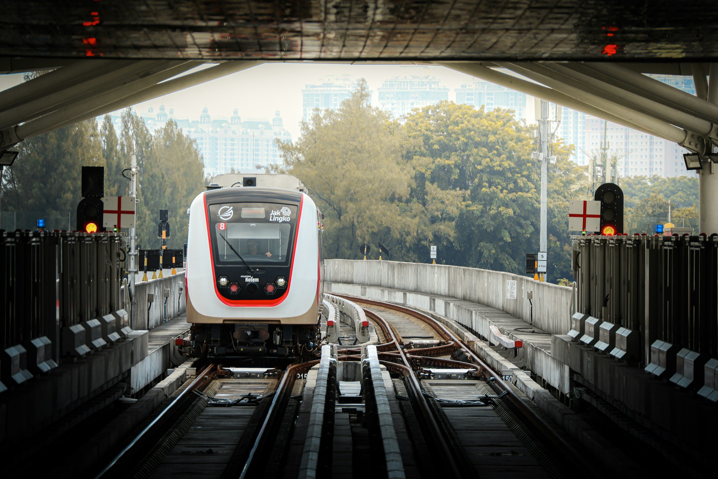 A train arriving at an underground station with tracks and safety barriers, with trees and buildings visible in the background.