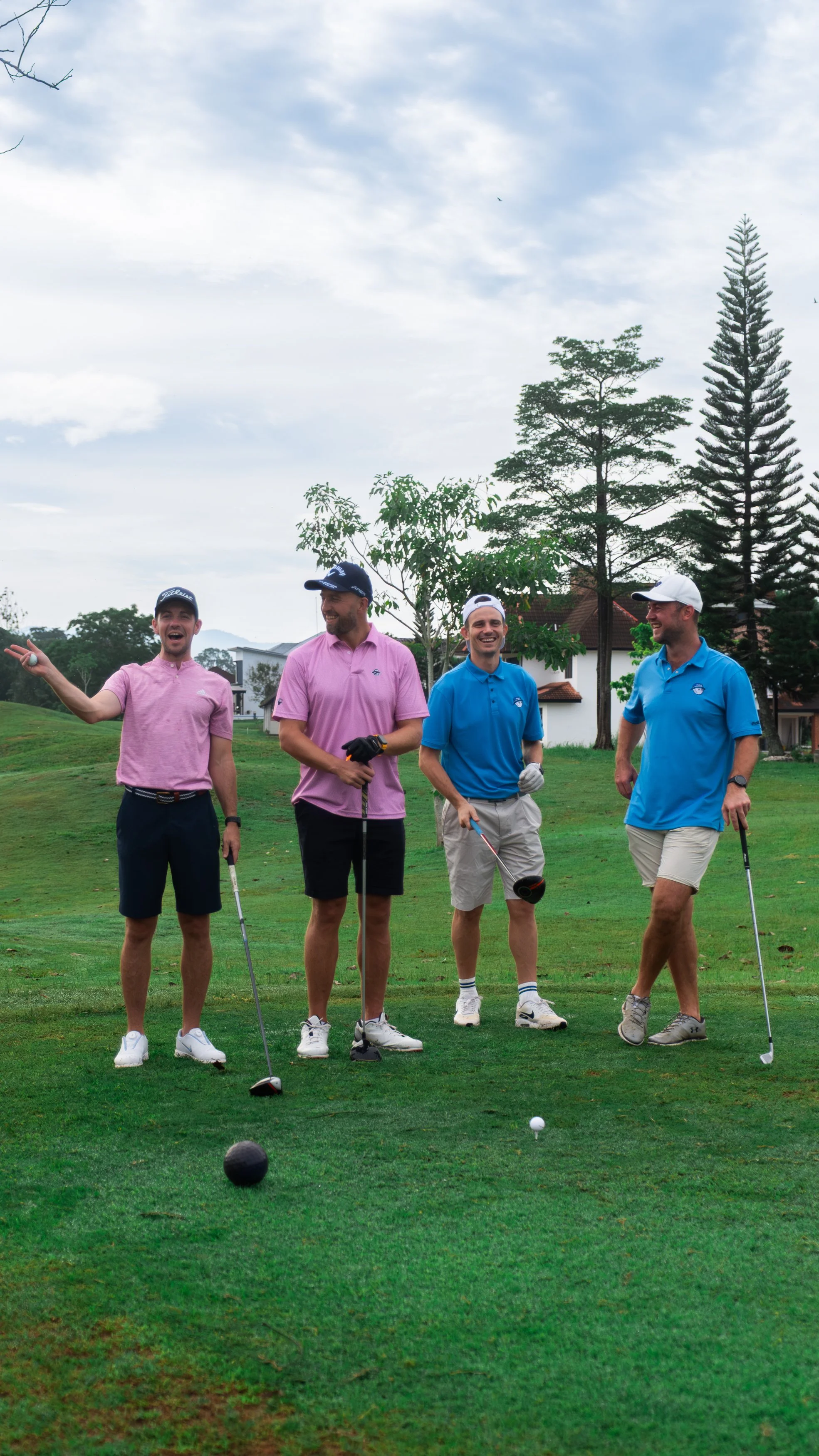 Four men on a golf course, wearing colorful shirts and shorts, enjoying a casual moment with golf clubs.