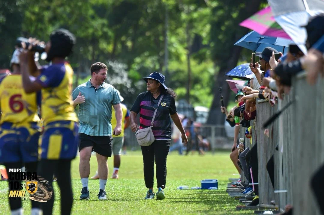 A woman and a man walking on a grassy field during a sports event, with spectators holding umbrellas on the side.
