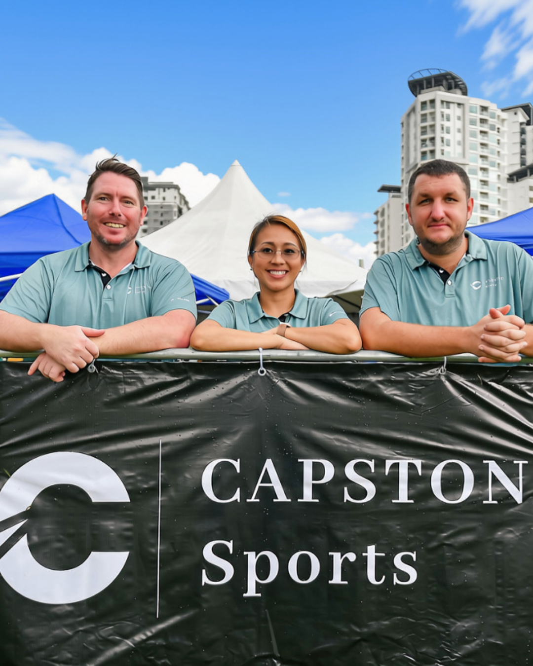 Three smiling individuals, two men and one woman, leaning on a barrier at a sports event, with tents and tall buildings in the background.