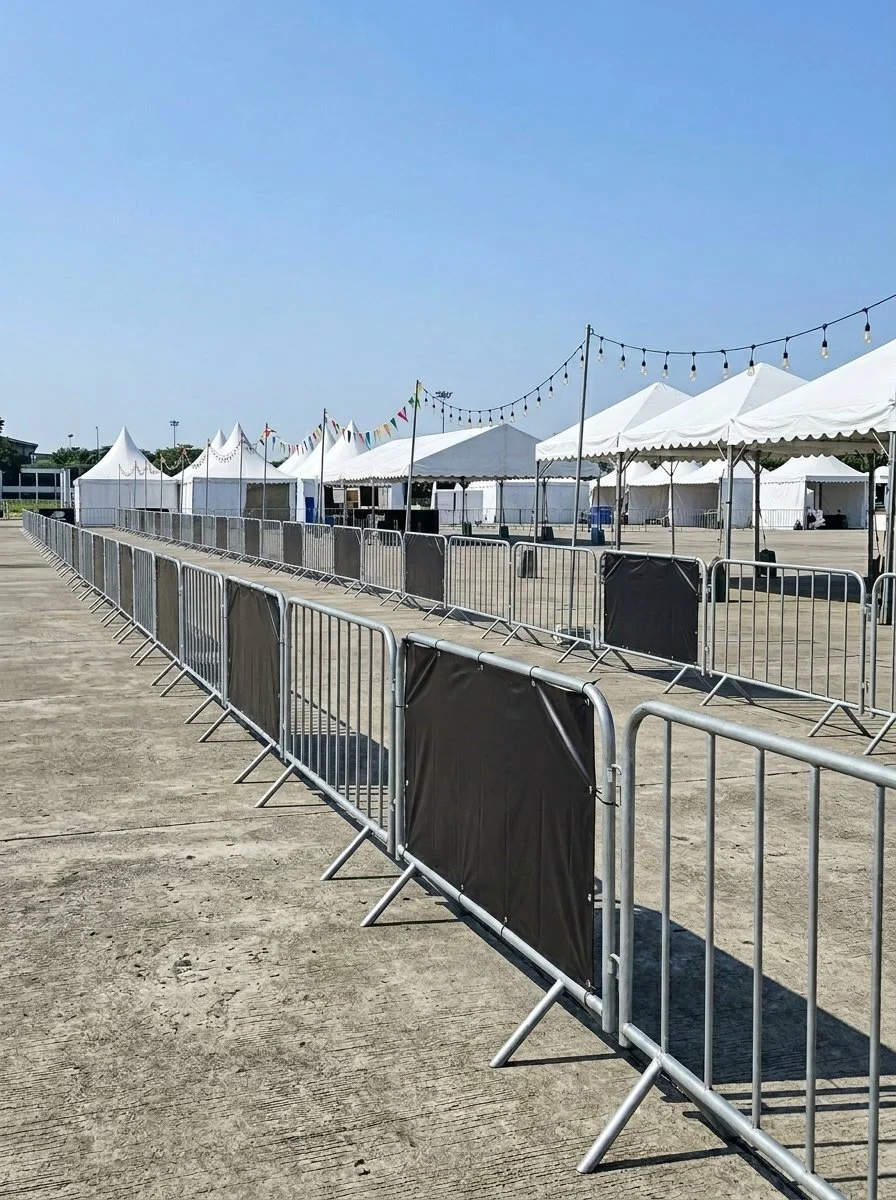 Empty event area with white tents, string lights, and metal barricades set up on a concrete surface under a clear blue sky.