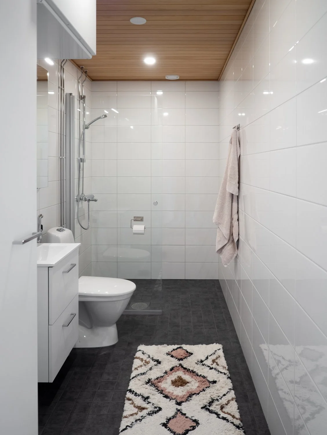 Modern bathroom with white tiled walls, gray floor tiles, a wooden ceiling, a wall-mounted shower, toilet, sink with cabinet, small vanity mirror, wall towel, and patterned rug.