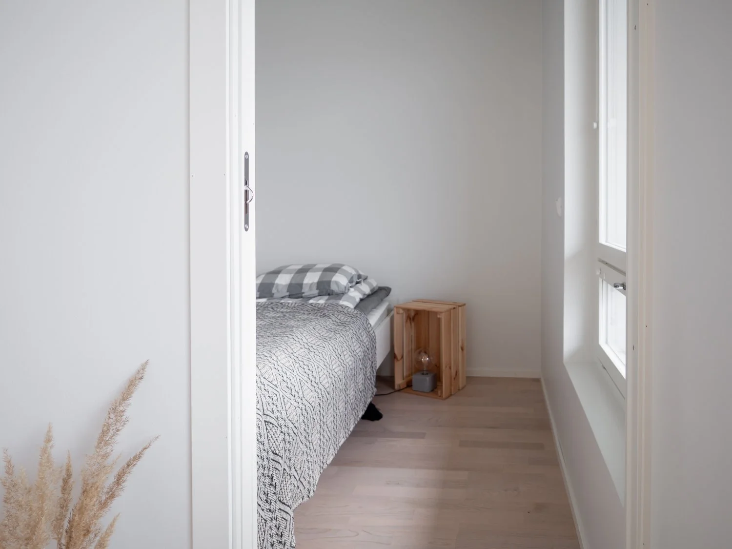 Minimalist bedroom with white walls, bed with checkered pillow and patterned blanket, wooden crate nightstand, window, and pampas grass decor.