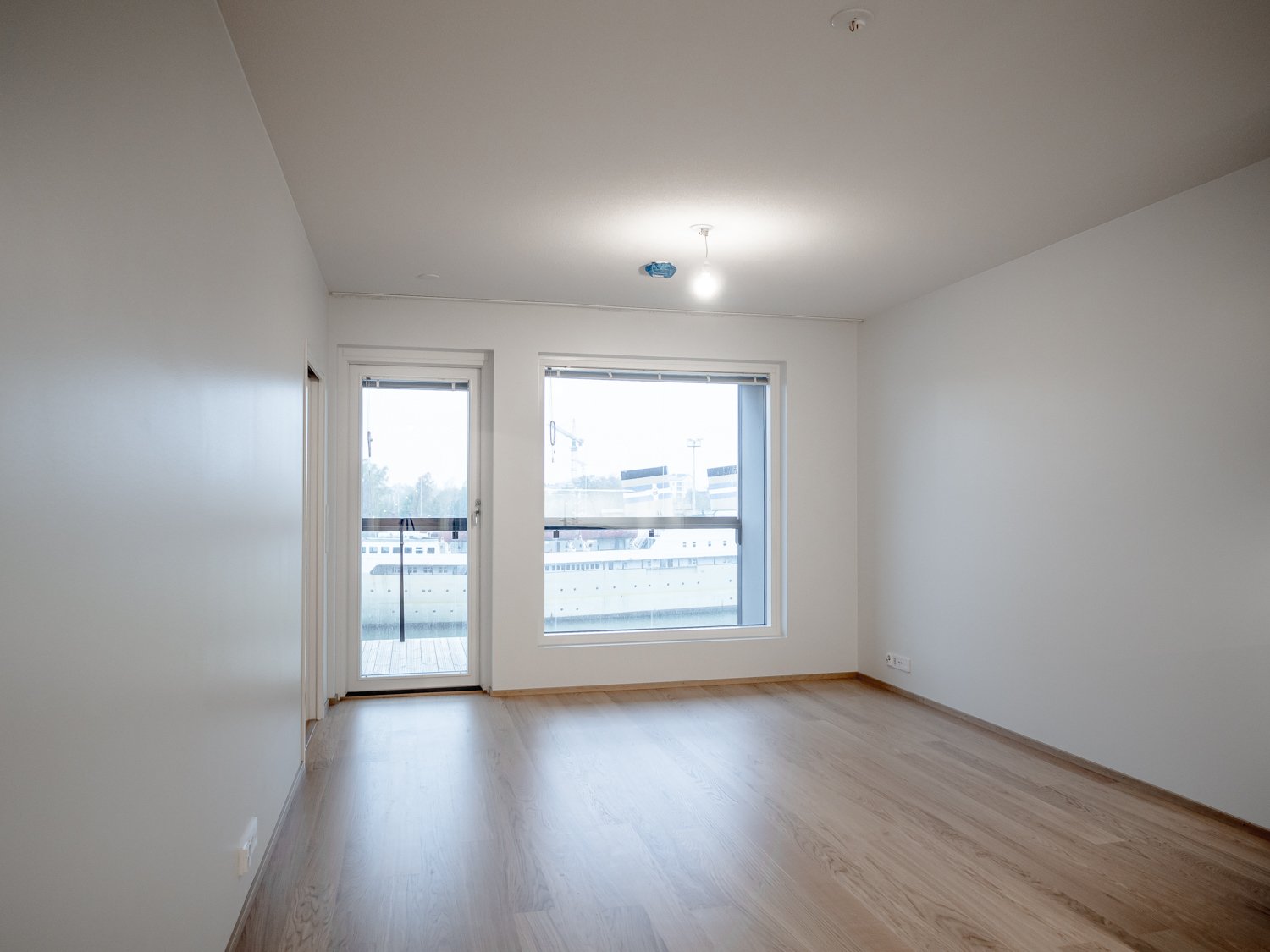 Empty room with wooden floor, large window, and glass door leading to a balcony.