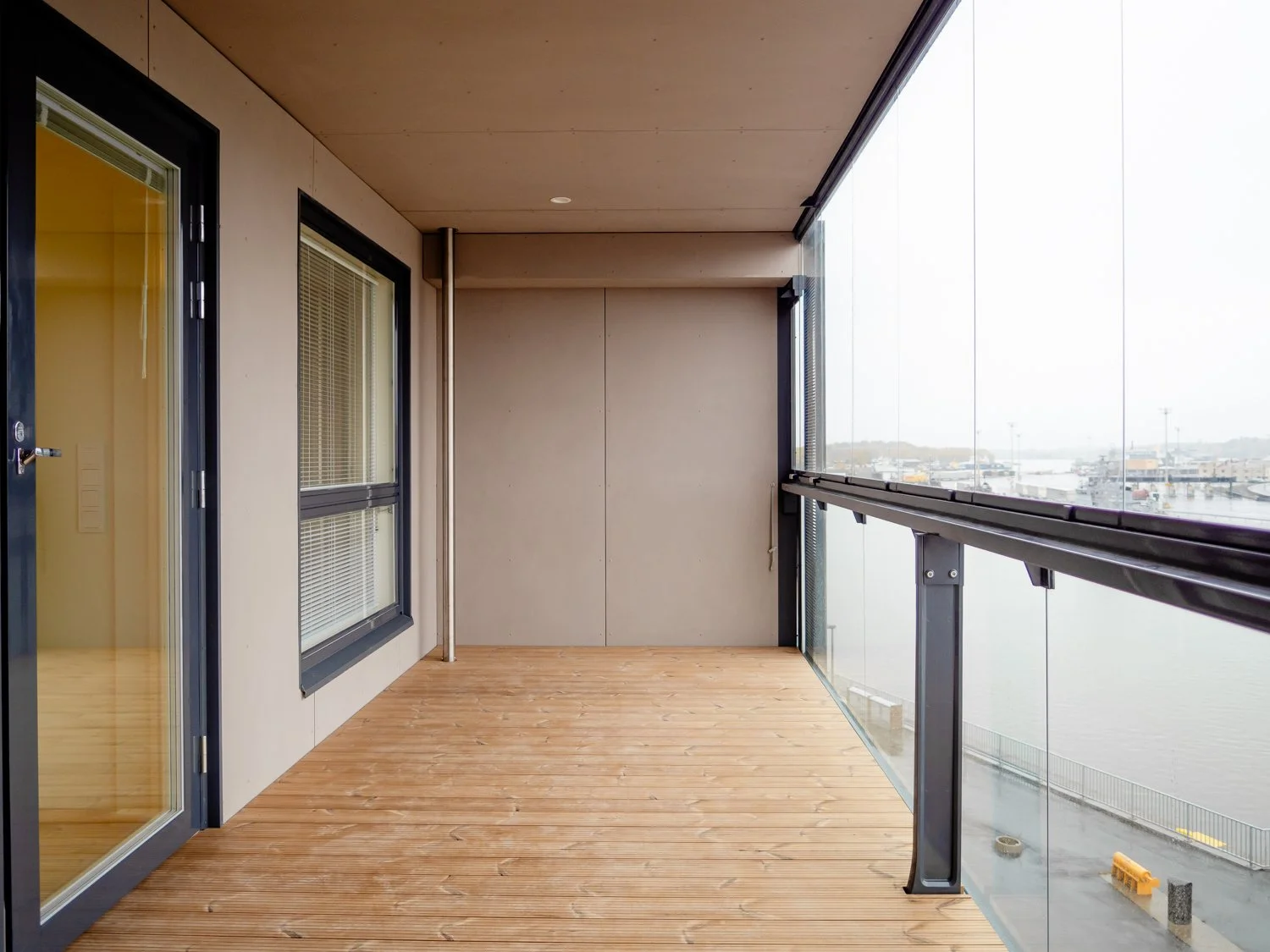 Modern enclosed balcony with glass railing overlooking a harbor; wooden floor and beige walls.