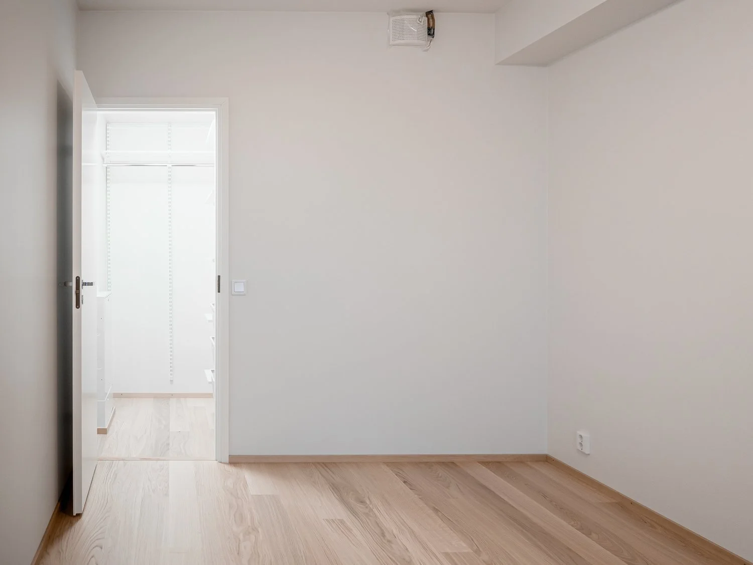 Empty room with light wood flooring, white walls, and an open door leading to a well-lit closet with shelves.